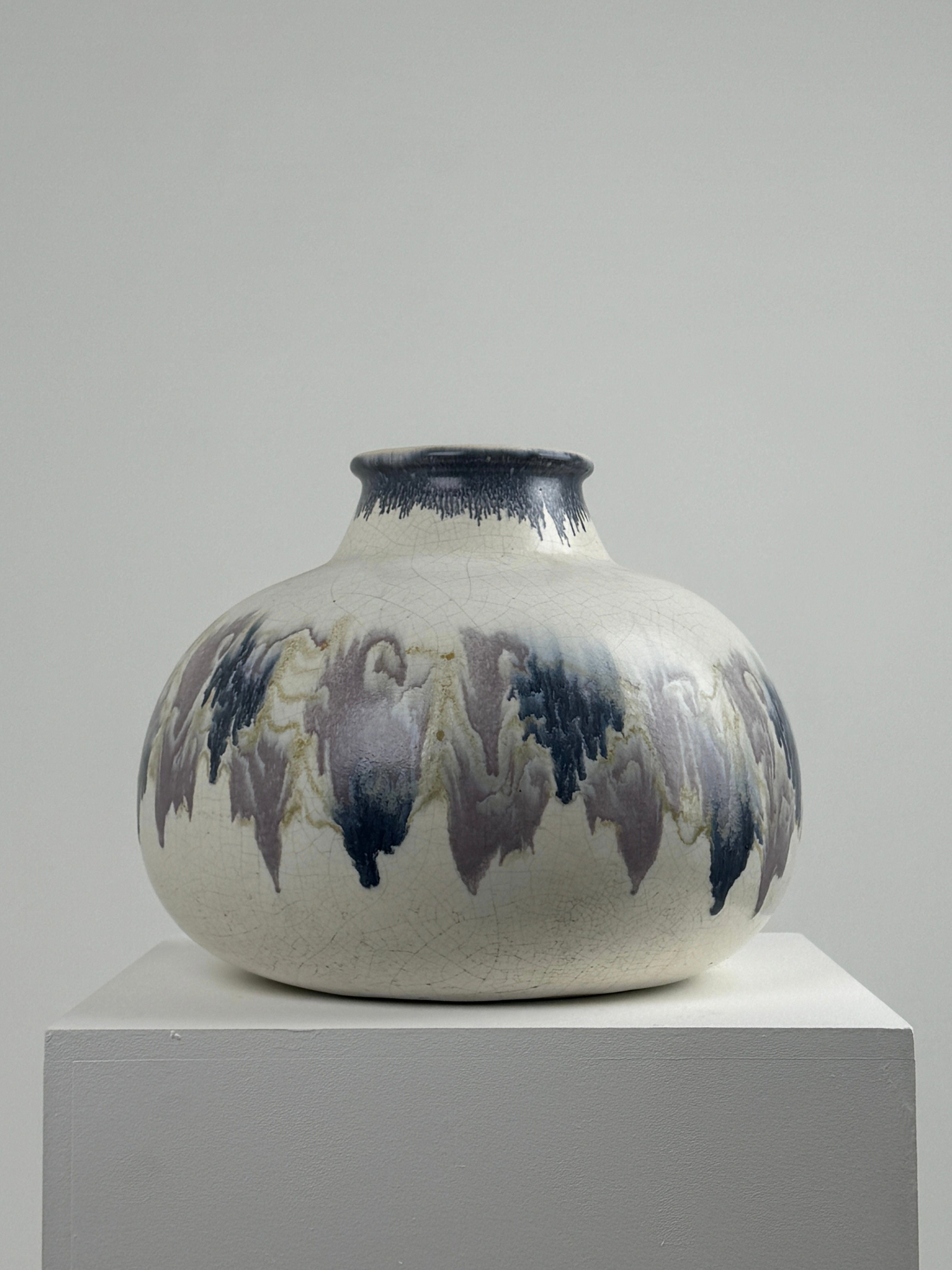 A Large ceramic vase by Ateliers Primavera from Figuresdesign is displayed on a white pedestal. The round vase, featuring a narrow opening and crackled glaze, boasts an Atelier Primavera-inspired surface with abstract wavy patterns in polychrome shades of blue, gray, and beige set against a neutral backdrop.