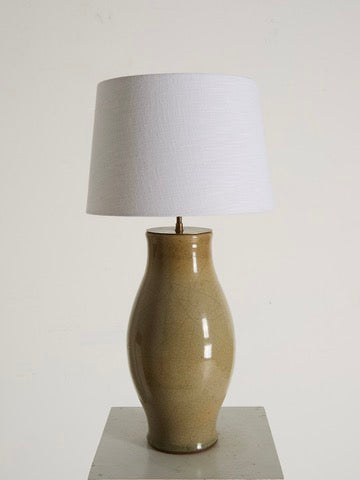 The 'Craquelure' Porcelain Lamp by Ancien et Jolie features a beige curvy base with a craquelure glaze and is topped with a white cylindrical shade. It rests on a light surface against a plain white background.