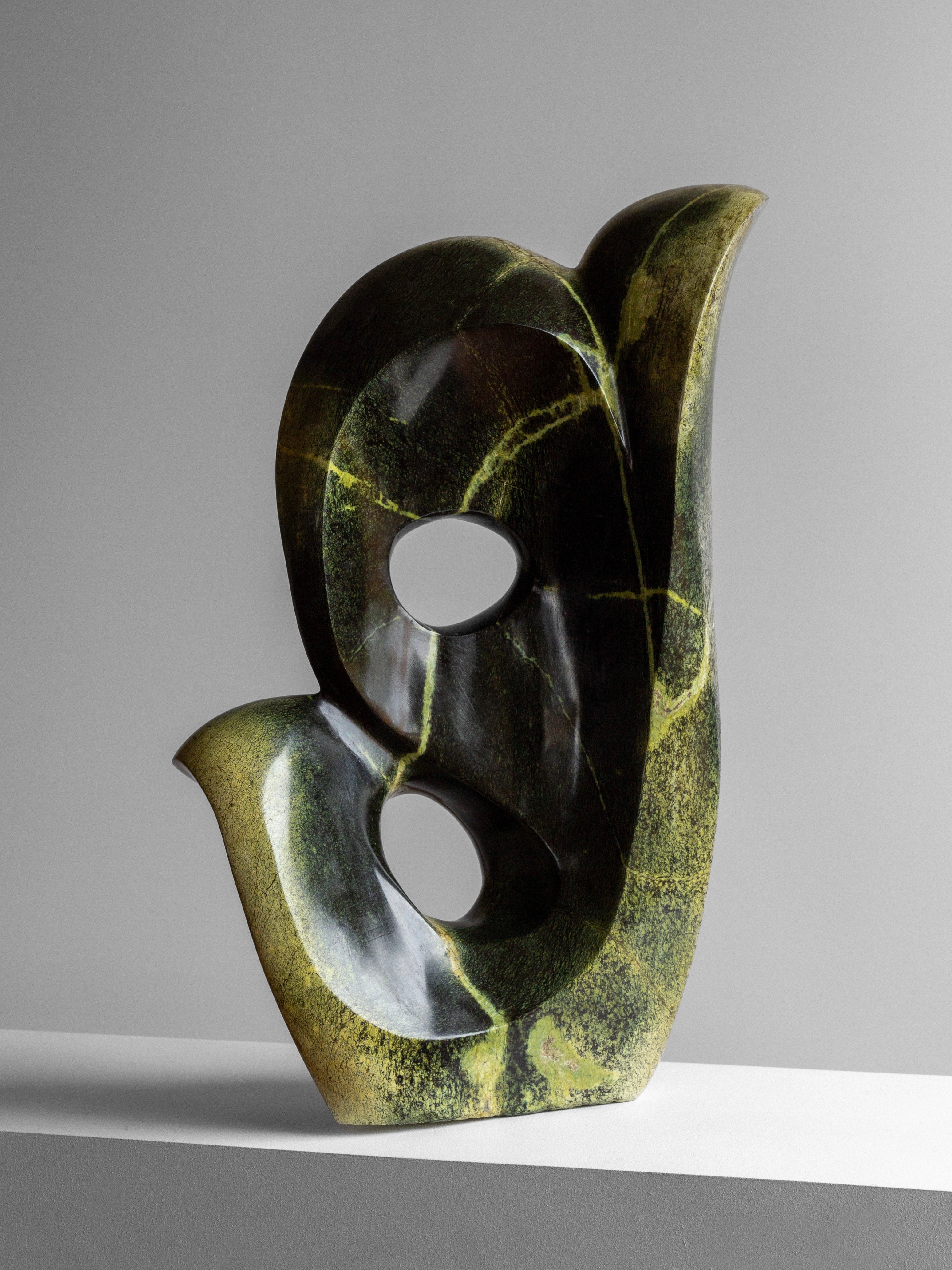 Abstract Stone Sculpture II by Spigel is a dark green sculpture with smooth curves and circular openings, crafted from leopard stone with lighter green veins. It boasts a glossy finish and rests on a white surface against a gray background.