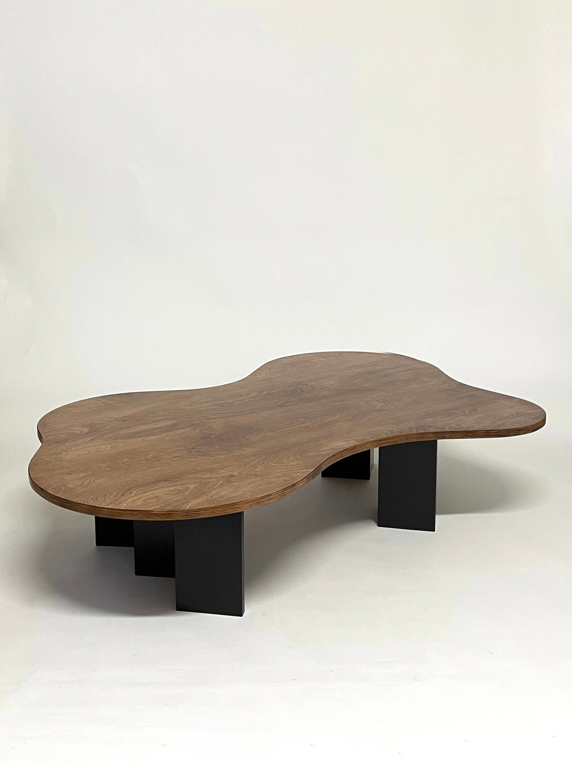 The Spill Table - Oak Black from GOONS showcases expert handcrafted details, boasting an irregular, curvy wooden top with sleek black rectangular legs, creating a striking contrast against a plain white background.