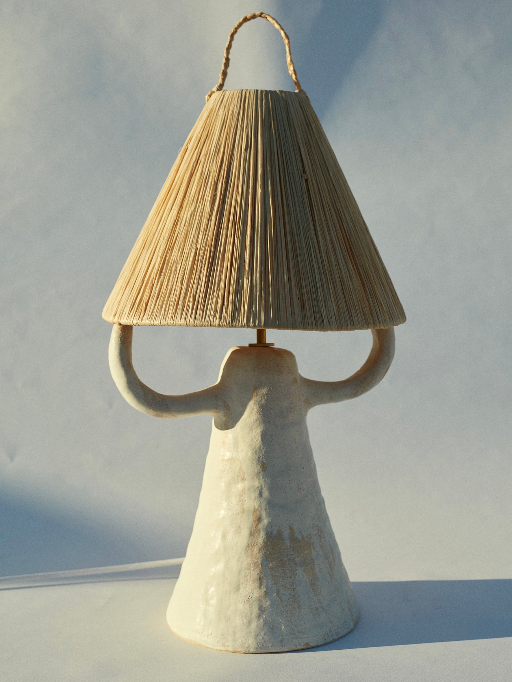 The Dona Table Lamp by marta bonilla features a textured, conical stoneware base with two curved handles, paired with a natural raffia shade for an earthy and artistic look against a plain backdrop.