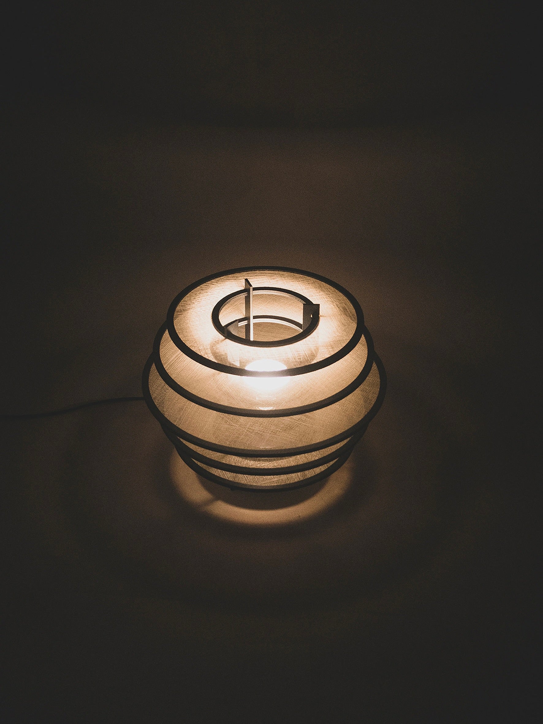 In a dimly lit space, ELIURPI's Lamp I—a softly glowing, sculptural piece with a spherical, layered design—sits gracefully. This handcrafted lamp emits warm ambient light that gently illuminates its surroundings, creating a cozy and serene atmosphere. A thin cord extends from the base of Lamp I.