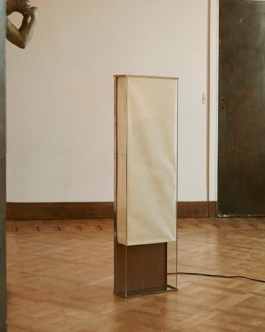 The Floor Lamp 2022 by Studio HAOS, featuring a tall rectangular minimalist design with a translucent cream shade and clear base, stands on a wooden parquet floor. A black power cord extends from its side; part of a sculpture appears on the left.