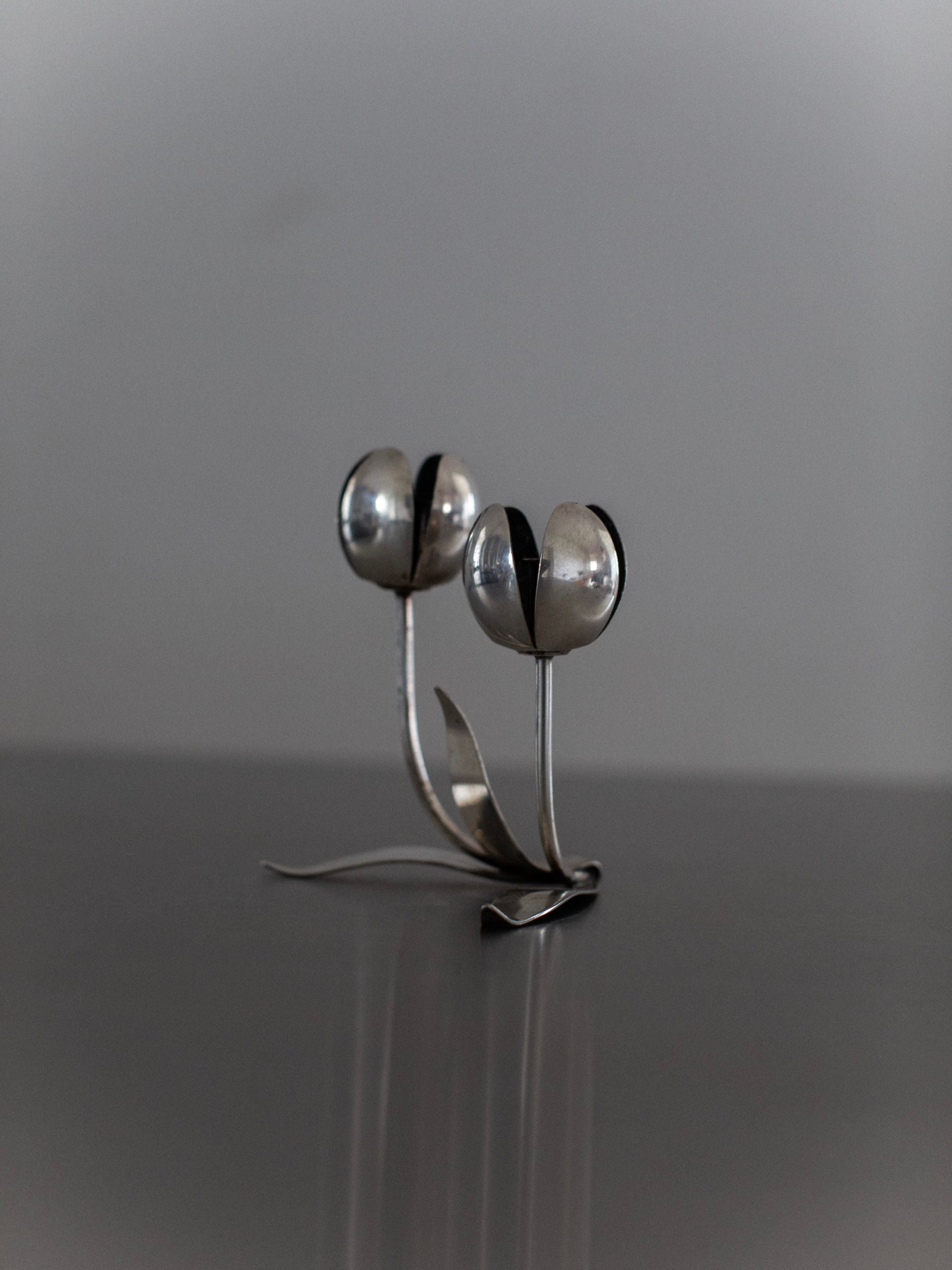 Against a plain gray backdrop, the Steel Tulip Candleholder by Out For Lunch adds a touch of modern elegance to any home decor. The silver sculpture, with tulip-shaped flowers featuring long stems and leaves, beautifully reflects light on its smooth metallic surface.