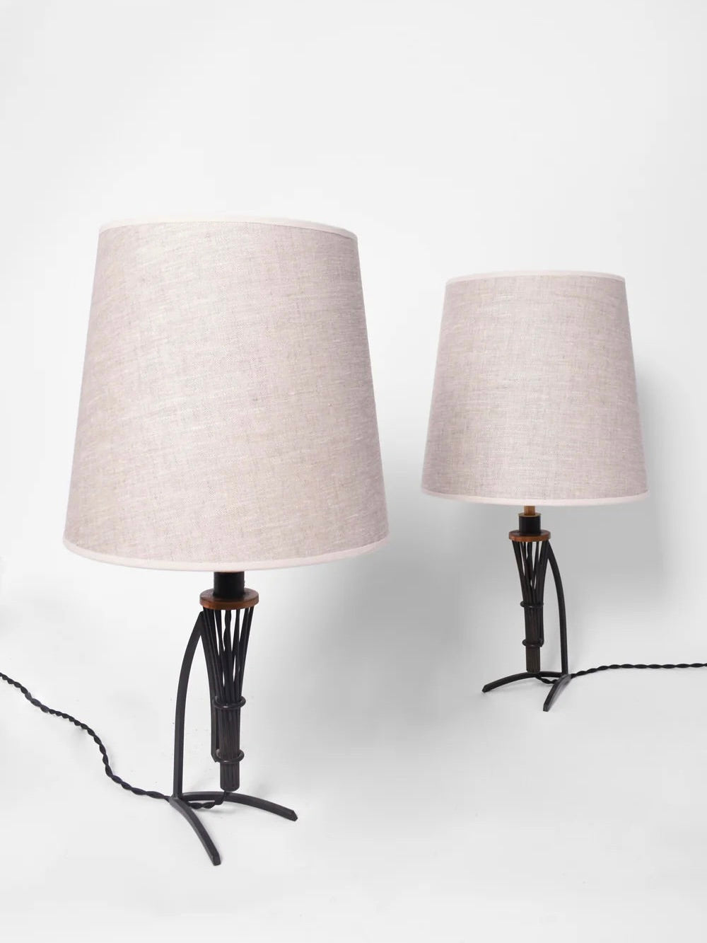 A pair of Barracuda Interiors' Iron And Copper Lamps, reminiscent of 1940s France, featuring a classic twisted design on the base. These black wrought iron lamps come with beige fabric lampshades and are connected by a sleek black braided cable, set against a white background.