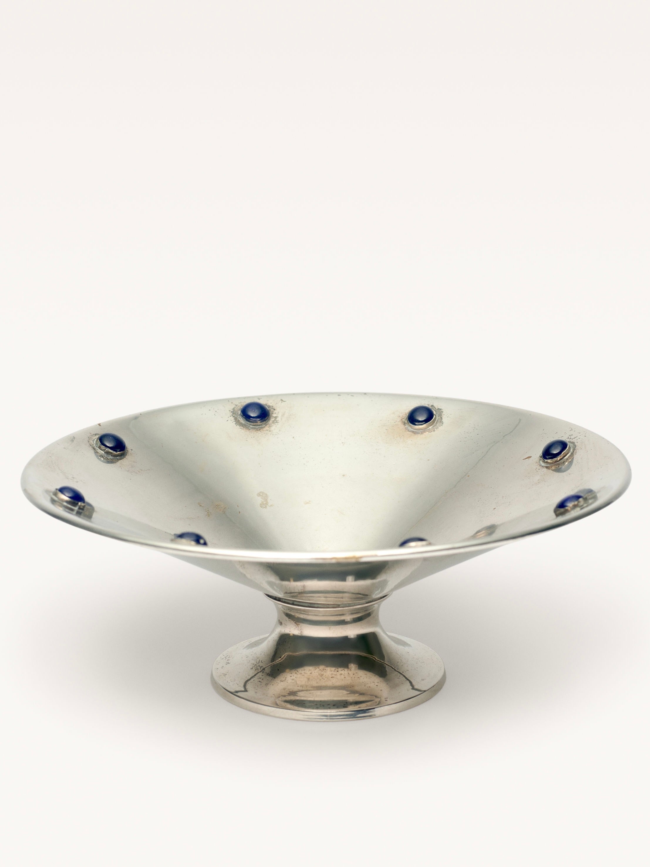 The "Silver Plated Bowl with Blue Stones" by Galerie MLS is a wide, shallow pedestal bowl featuring blue stones around the rim. It has a reflective surface, stands on a rounded base, and shows slight signs of use that enhance its elegance.