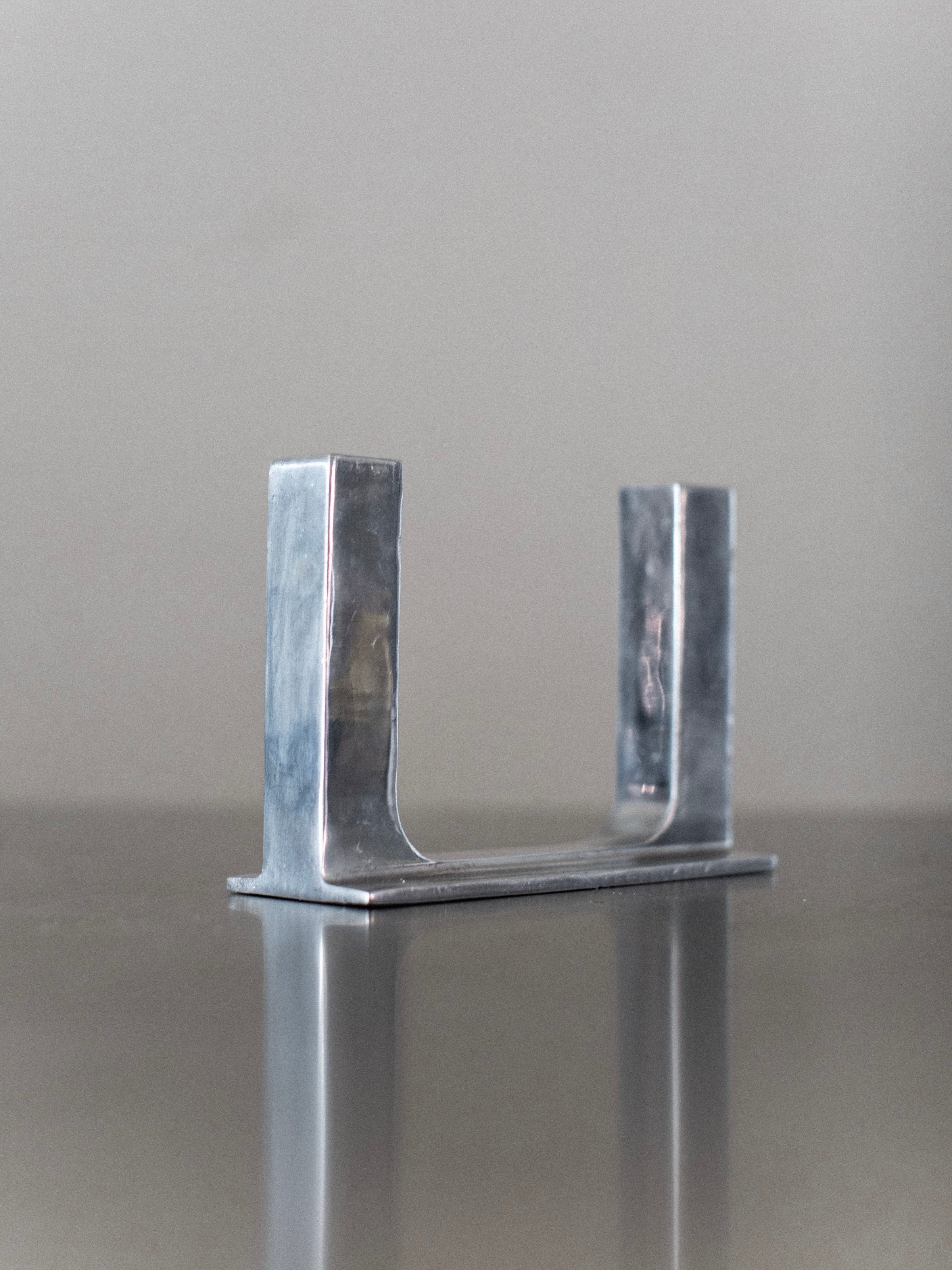 The Chrome Candleholder by Out For Lunch, a minimalist U-shaped steel piece with a subtle polished silver finish, is placed on a reflective surface. A plain, muted background allows this elegant object to take center stage, its metal catching the light with a gentle shine.