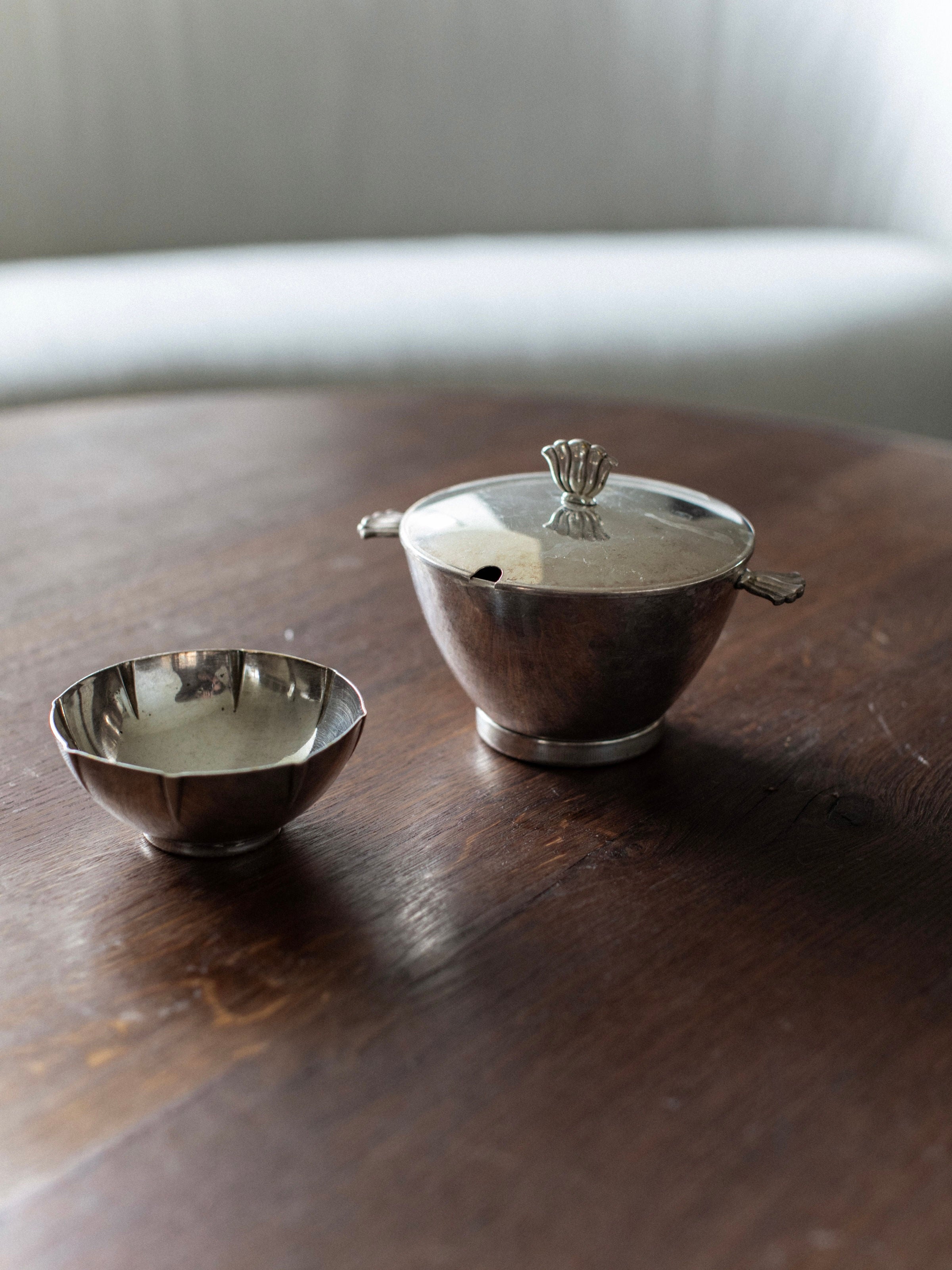 The Art Deco Bowls from Out For Lunch exude vintage glamour as they sit elegantly on a wooden table, featuring a small silver bowl and a lidded silver sugar bowl with ornate handles. In the background, a blurred, light-colored couch adds to the timeless charm of this unique natural story.
