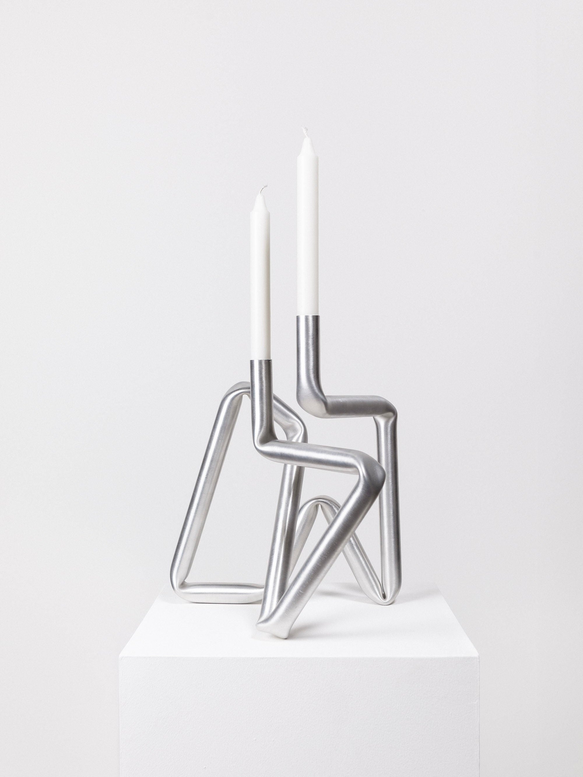An AOT STUDIO Bucati Candle Holder (brushed aluminium) with an abstract geometric design featuring interconnected aluminium tubes holds two white taper candles. This hand-made modern piece is displayed on a white pedestal against a plain white background.
