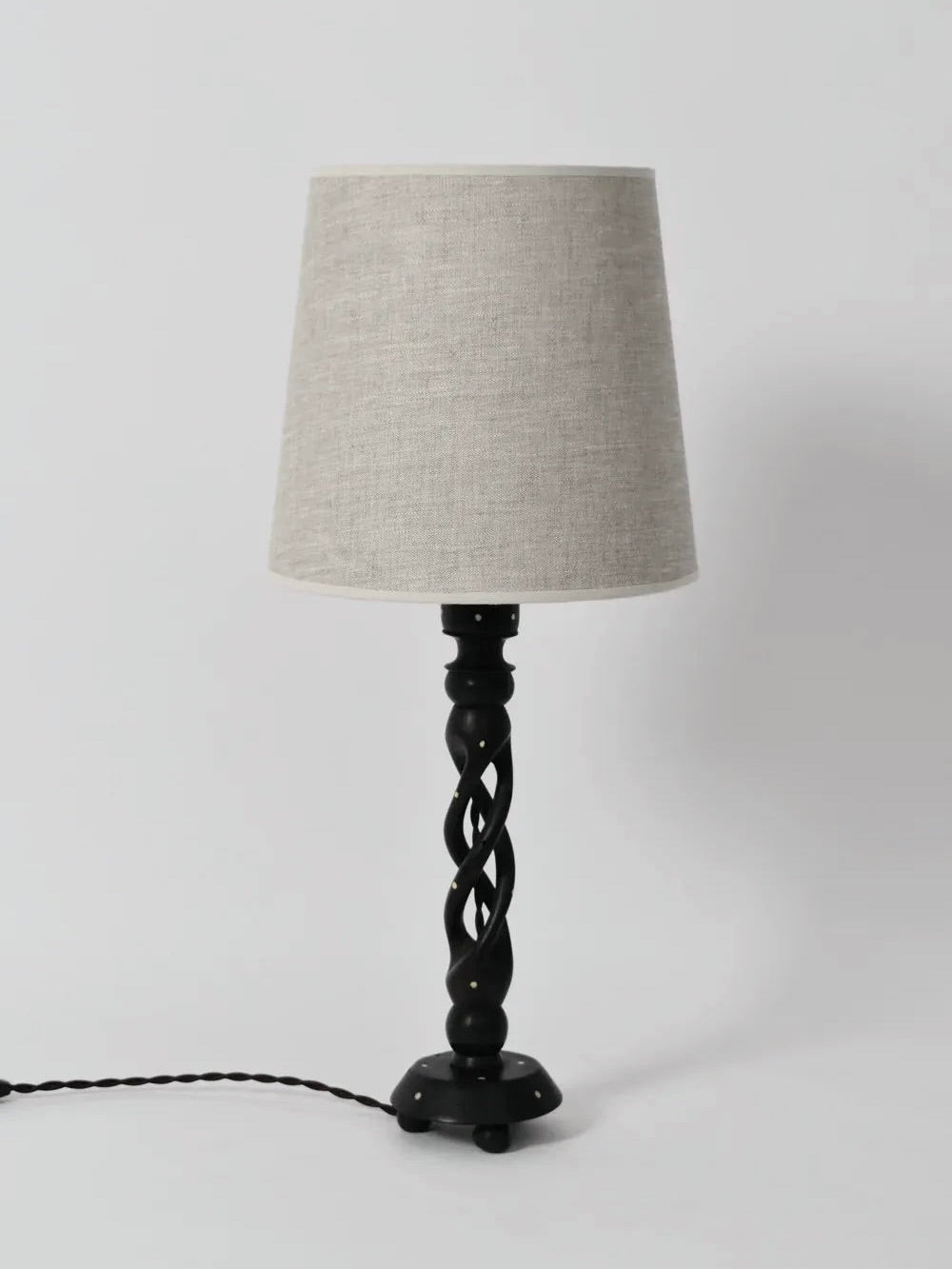 A black table lamp, known as the Turned Wood Twist Lamp from the 1960s by Barracuda Interiors, features an elegant twisted ebonised wood base complemented by a beige fabric lampshade. The lamp's power cord is visible against a plain white backdrop.