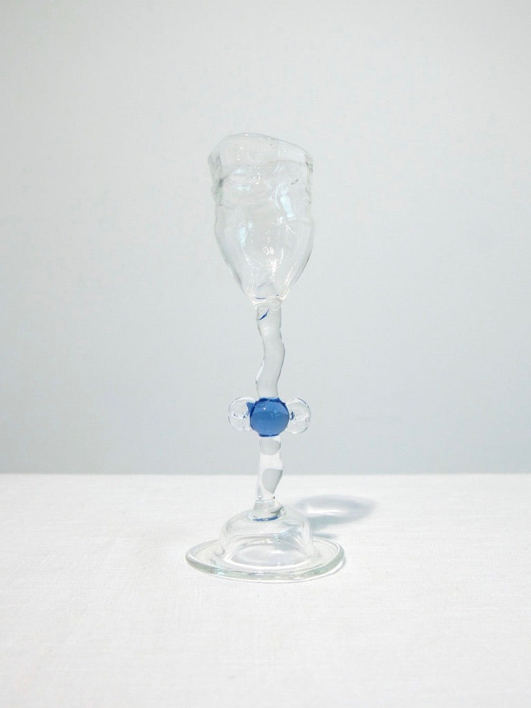 The Vivian Alarcón Blue 'Candy' Mouth Blown Shot Glass, featuring a twisted stem and blue glass bead at the center, rests on a white surface against a light background.