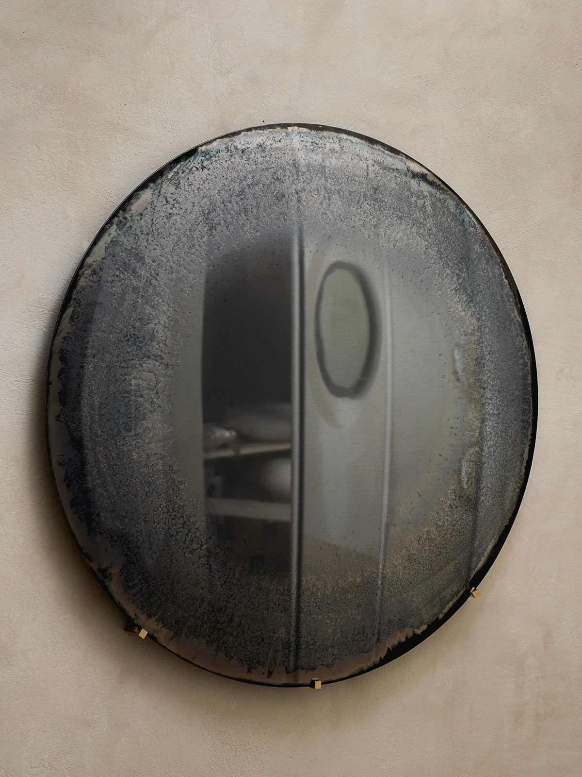 The Gray Mist Convex Oxidized Mirror by HOMA casts a hazy reflection of a contemporary white bathroom, featuring an oval mirror and countertop. This smoke-colored piece elegantly hangs on a textured neutral wall with its brass hanger.
