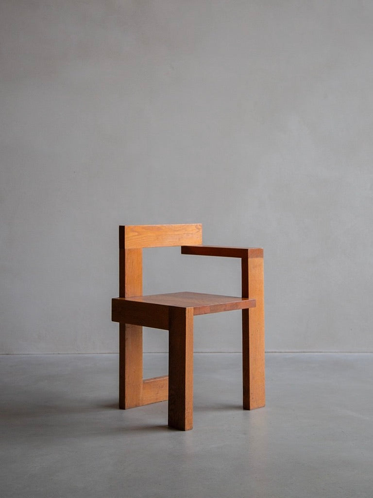 A minimalist wooden chair, the Manner Of Gerrit Rietveld by Bicci de' Medici, features a rectangular backrest and armrests. This mid-century modern piece with simple geometric lines sits elegantly on a concrete floor against a plain gray wall in a natural solid oak finish.