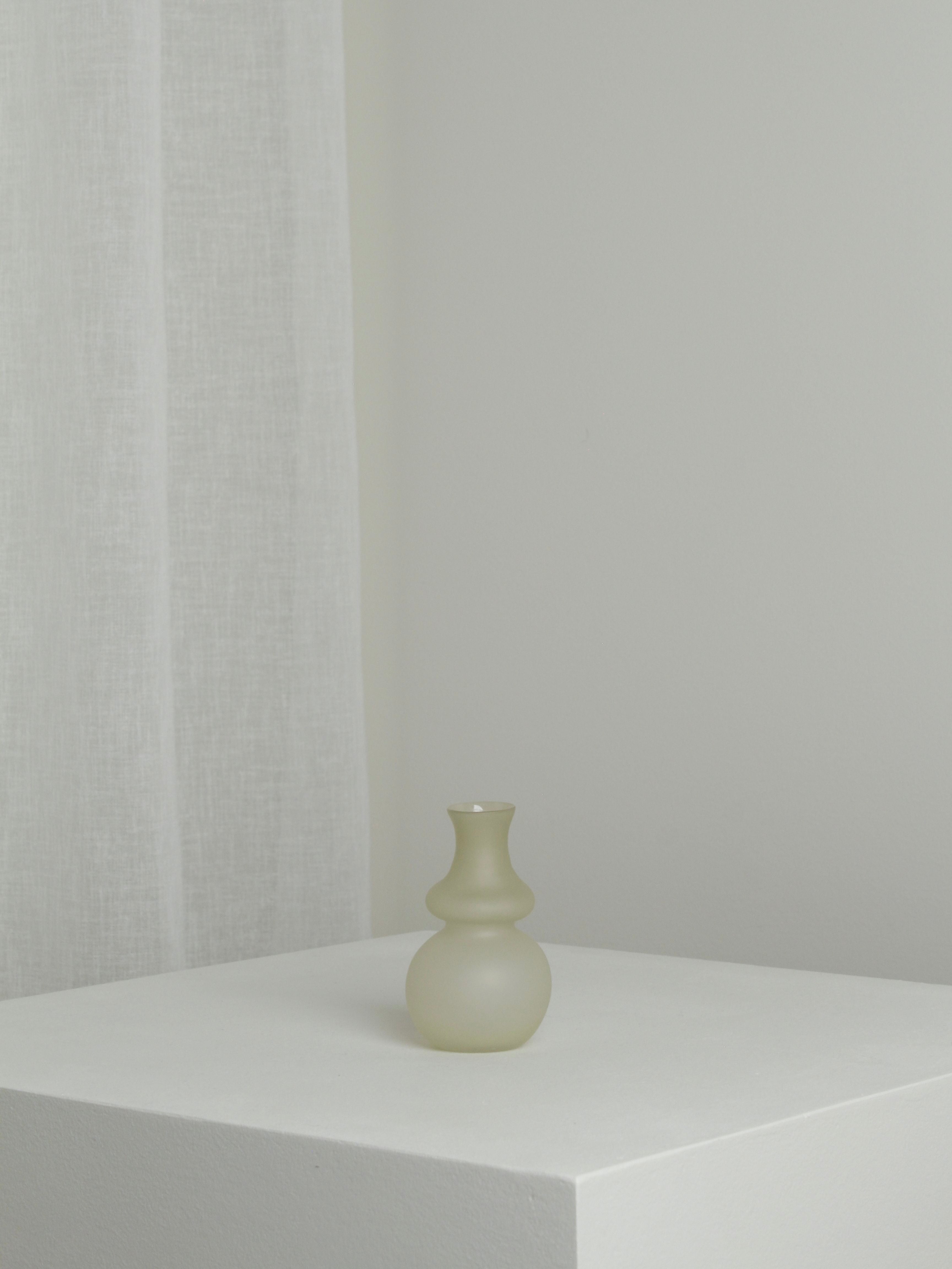 A Small Vase in Stone by Verre d'Onge, made of frosted glass with an elegant curvy design, sits like a cherished stone on a white pedestal in a minimalist setting. The background features an off-white wall and a sheer, white curtain. The overall aesthetic is simple and serene, reminiscent of a quiet corner in Montreal.