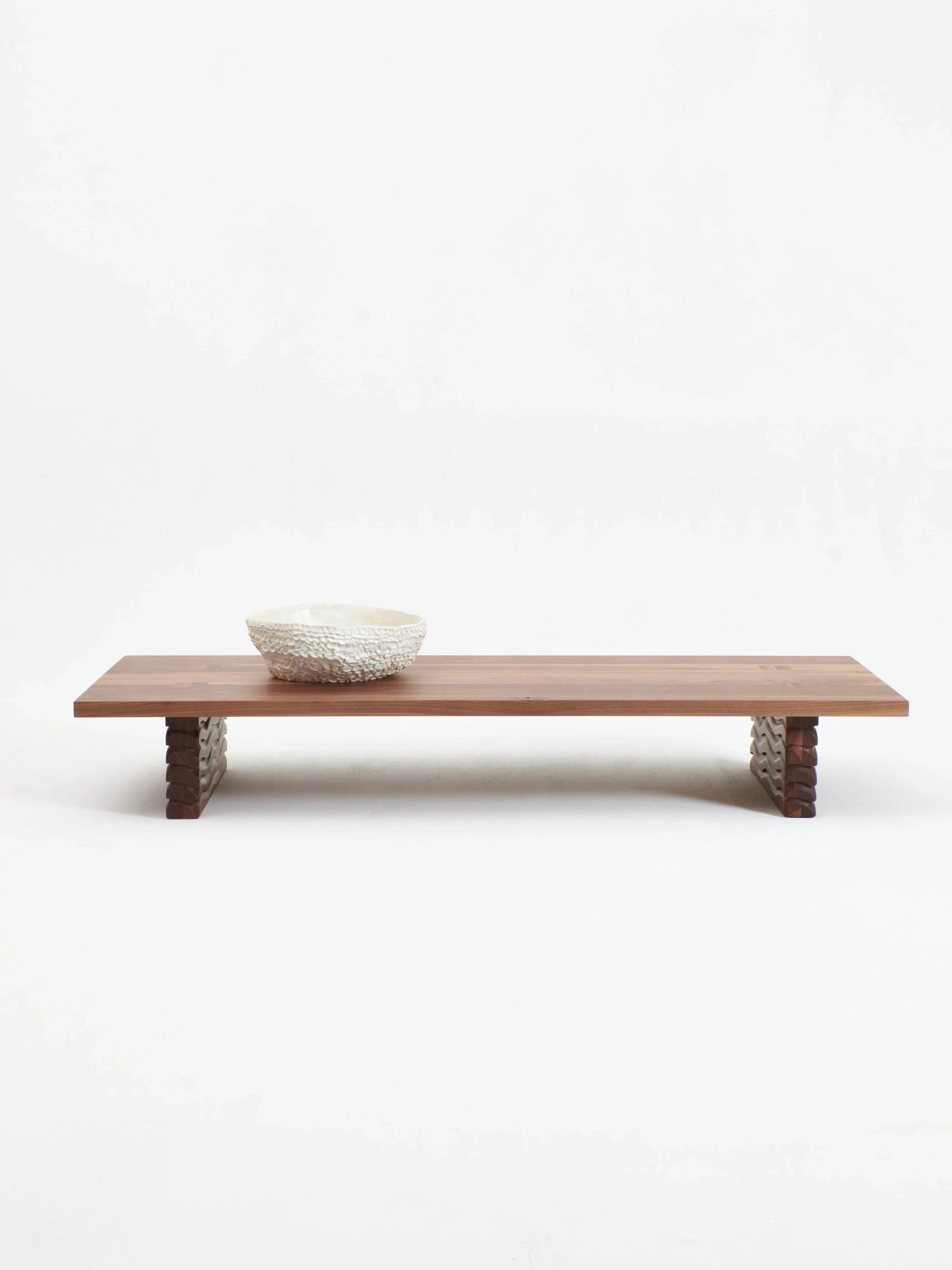 The Nazaré Coffee Table by Project 213A is a minimalist wooden masterpiece featuring intricate leg designs, handcrafted from solid walnut. It sits on a white background with a textured, shallow ceramic bowl on top, creating an effortlessly elegant aesthetic.