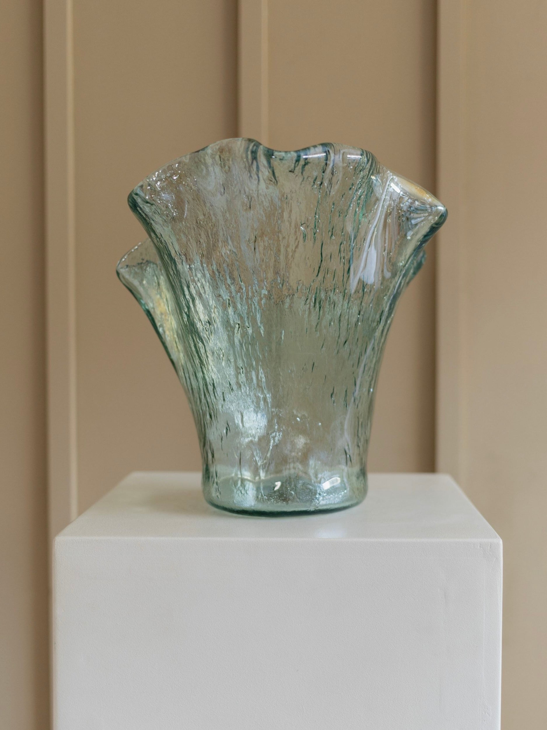 A Solid Vintage Murero Glass Vase by Bottega Jacobs, crafted in Italy during the 1970s, graces a white pedestal. Its wavy, fan-like shape and translucent, textured surface reveal subtle shades of green and blue against a beige paneled wall backdrop.