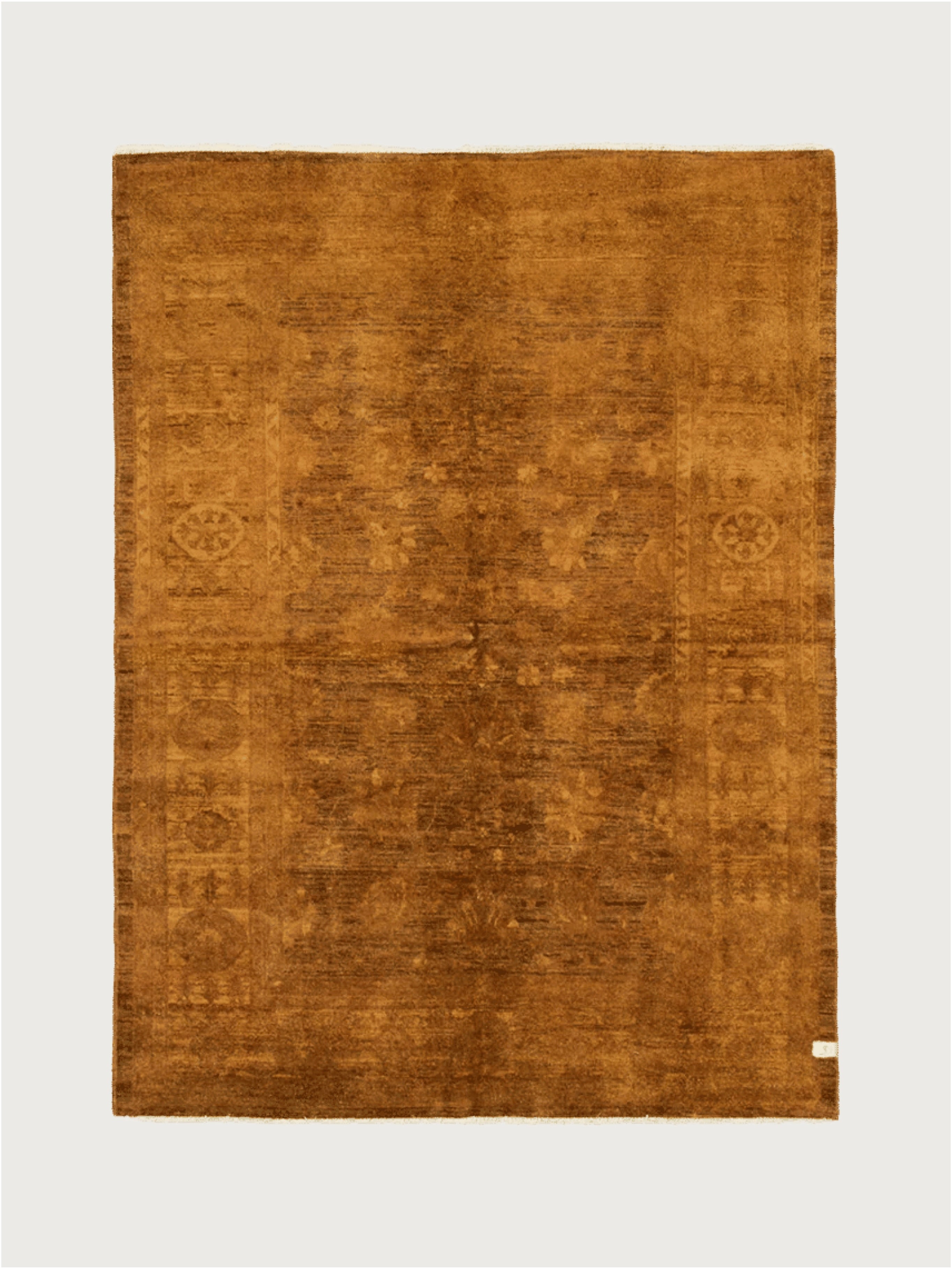 A square, hand-knotted Overdyed Rug in Safron by catalog:three, 193 x 261 cm, featuring a rich brown hue with textured wool patterns on a light neutral background and a small corner tag.