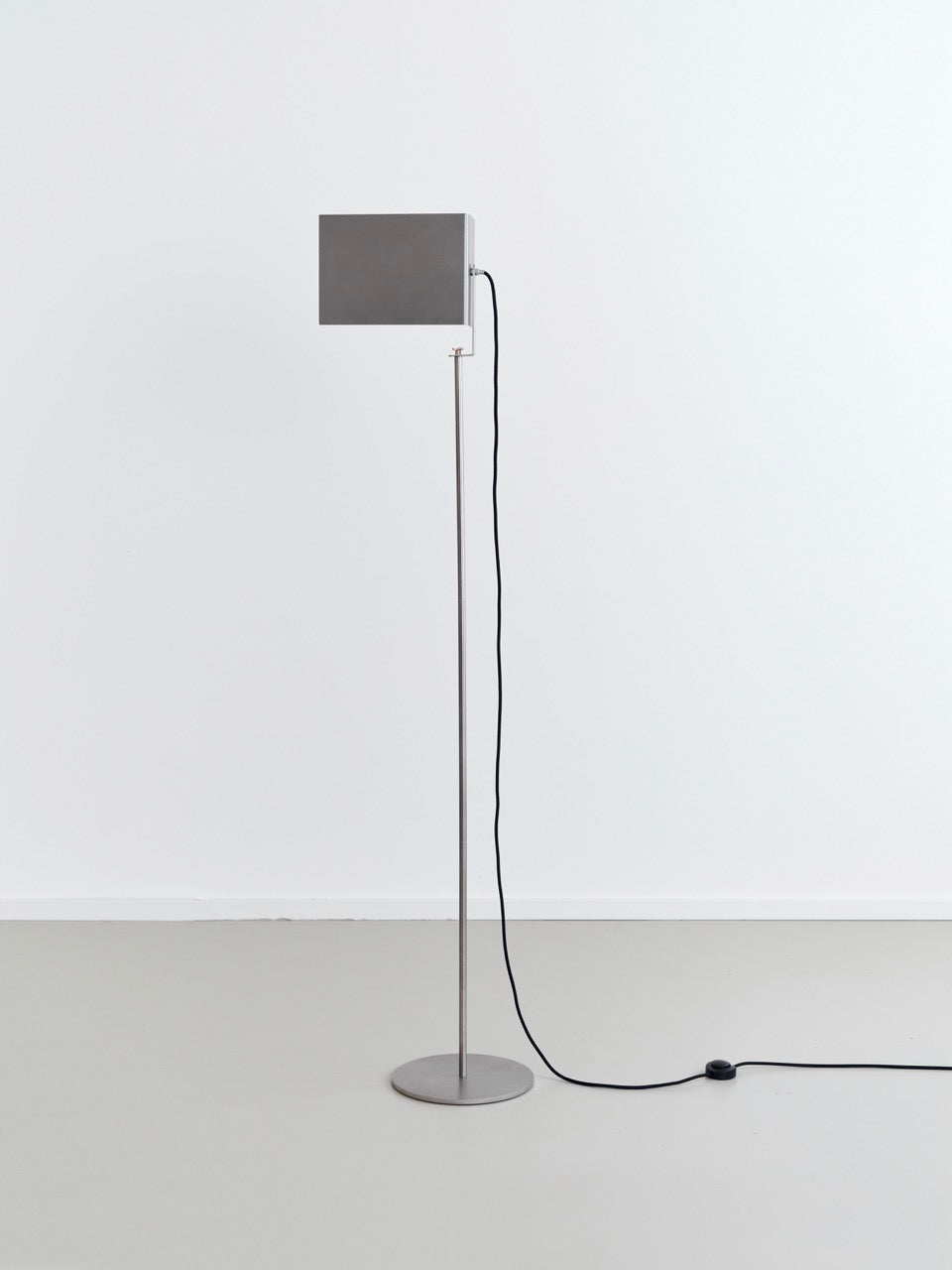 The "Flagpole" Floor Lamp by Frederik Fialin embodies functional simplicity with its minimalist design, featuring a rectangular gray shade atop a slender metallic stand. It includes a black power cord and foot switch, set against a plain white background on a light gray floor, reflecting the design principles reminiscent of Joseph Beuys.
