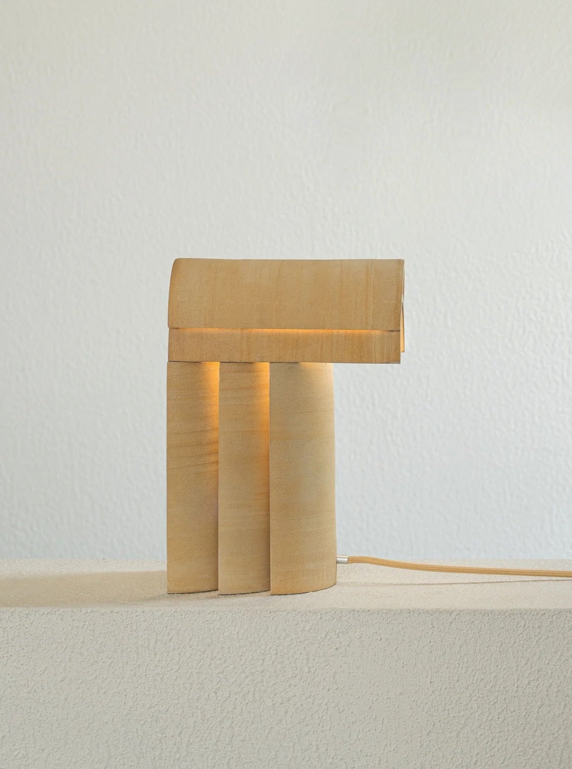 A minimalist sculptural lighting piece stands on a white surface against a light-colored wall. The Evelina Kudabaite "Layers of Light I" features a simple, cylindrical base supporting a rectangular shade, casting a warm glow.