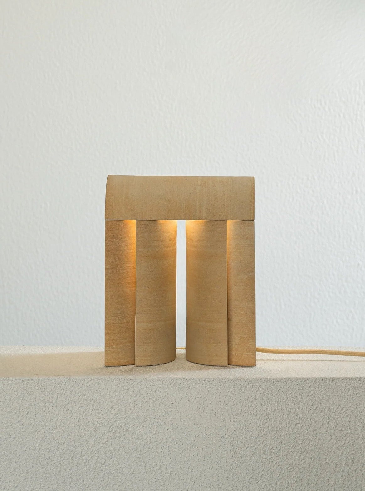 The "Layers of..." Light III by Evelina Kudabaite, a sculptural handcrafted wooden lamp with a minimalist arch-like design, sits on a textured white surface. It emits a soft glow, casting an elegant and warm ambiance against the plain wall backdrop.