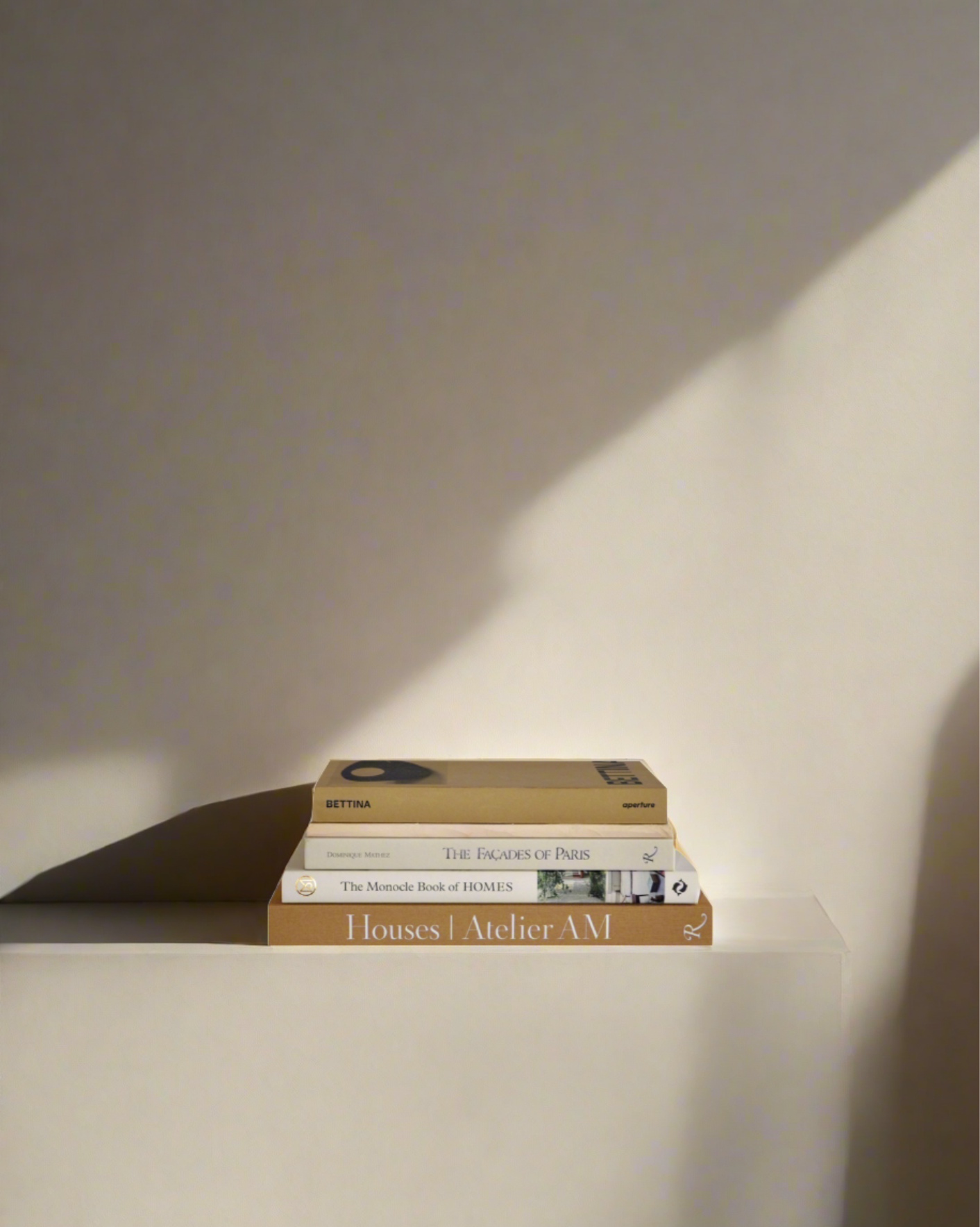 A stack of four art and design books from Maison Plage's Beige Set of 5 Art and Design Books rests on a white surface illuminated by soft sunlight. The visible titles are: "Houses Atelier AM" on the bottom, "The Monocle Book of Homes", "The Eames of Paris" in the middle, and "Fredun" on the top. Shadows cast on the wall accentuate these beautiful interiors reads.