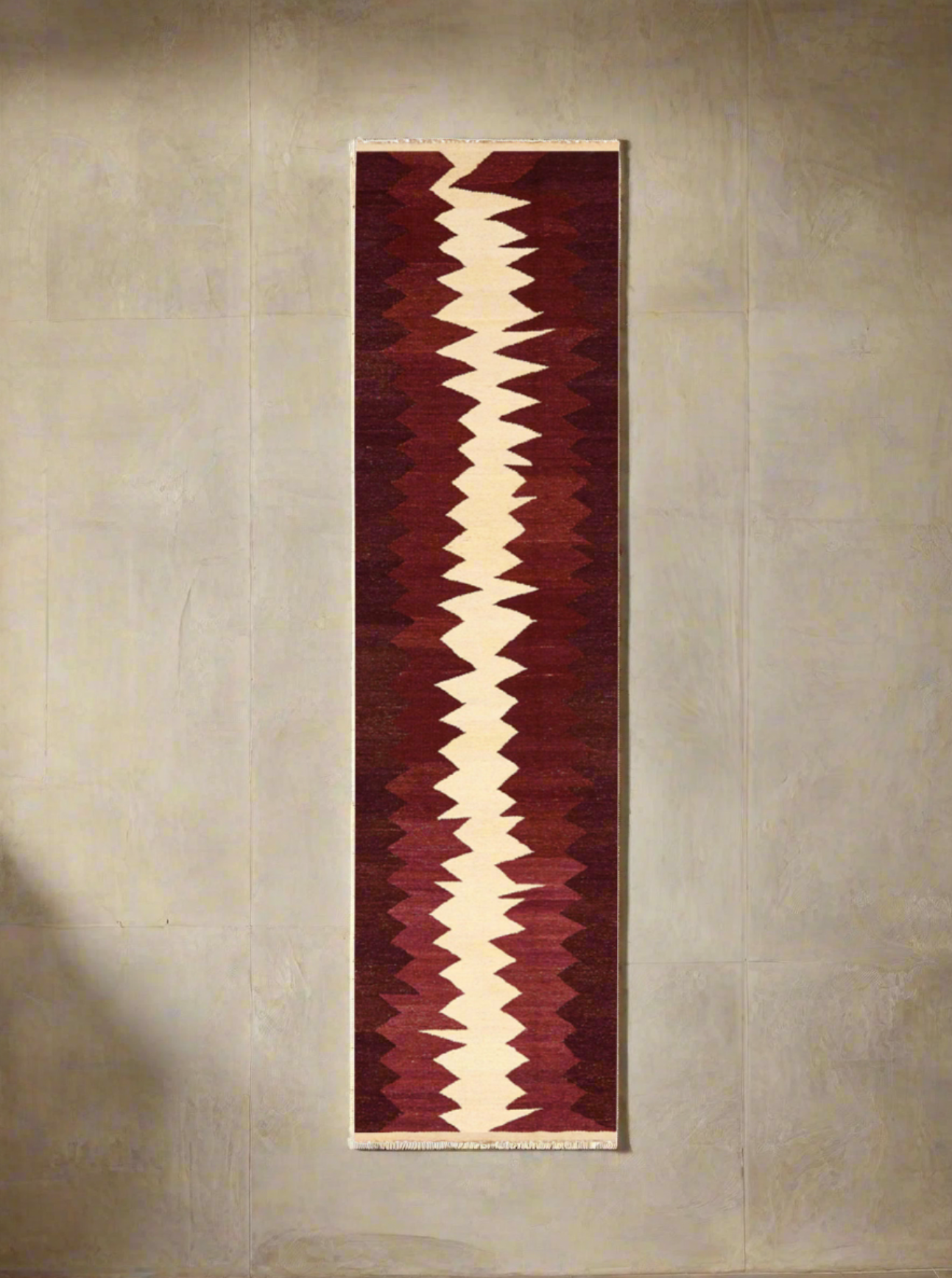 A long, narrow Zig Zag Kilim Runner - Handwoven Rug with a bold, zigzag pattern running down its center. The handwoven wool features alternating shades of cream and burgundy, set against a deep red background. Crafted with natural dyes, the rug boasts a geometric, tribal-inspired design. This exquisite piece is brought to you by ISHKAR.