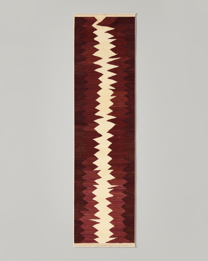 A long, narrow Zig Zag Kilim Runner - Handwoven Rug with a bold, zigzag pattern running down its center. The handwoven wool features alternating shades of cream and burgundy, set against a deep red background. Crafted with natural dyes, the rug boasts a geometric, tribal-inspired design. This exquisite piece is brought to you by ISHKAR.