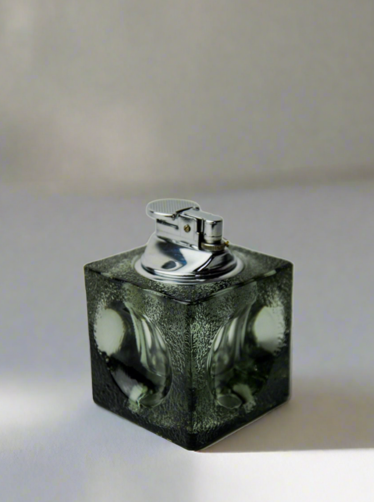 An Ice Cube Lighter by Antonio Imperatore from Malamar Studio, featuring a silver flip-top design, rests on a transparent, textured black Murano glass cube base with circular indents on each side. The background is plain and off-white.