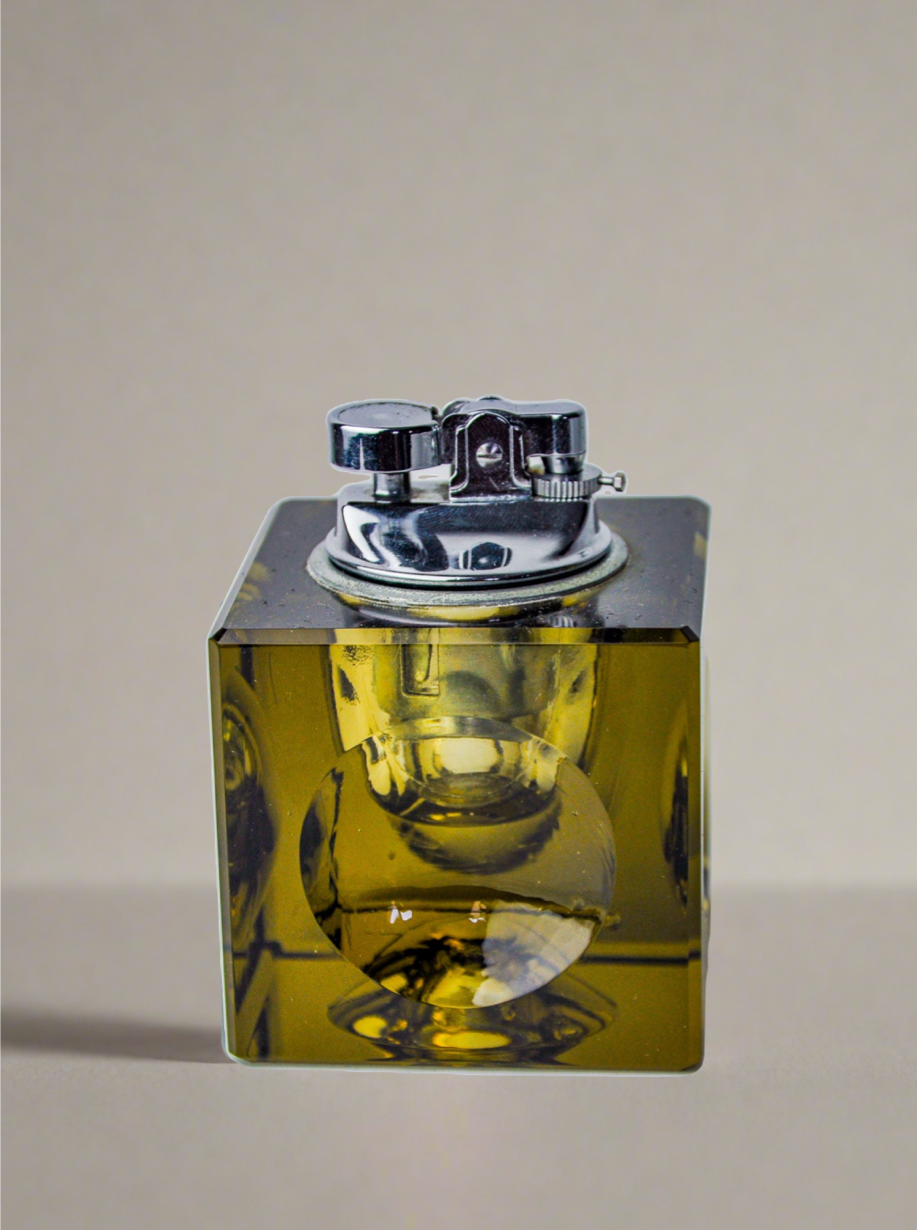 An exquisite Ice Cube Lighter by Antonio Imperatore, crafted by Venetian master glassmakers at Malamar Studio, rests elegantly on a flat surface. The vintage tabletop lighter features a reflective metal top and an intricate ignition mechanism, all encased in a transparent amber-colored Murano glass base that showcases its shiny finish against the plain background.