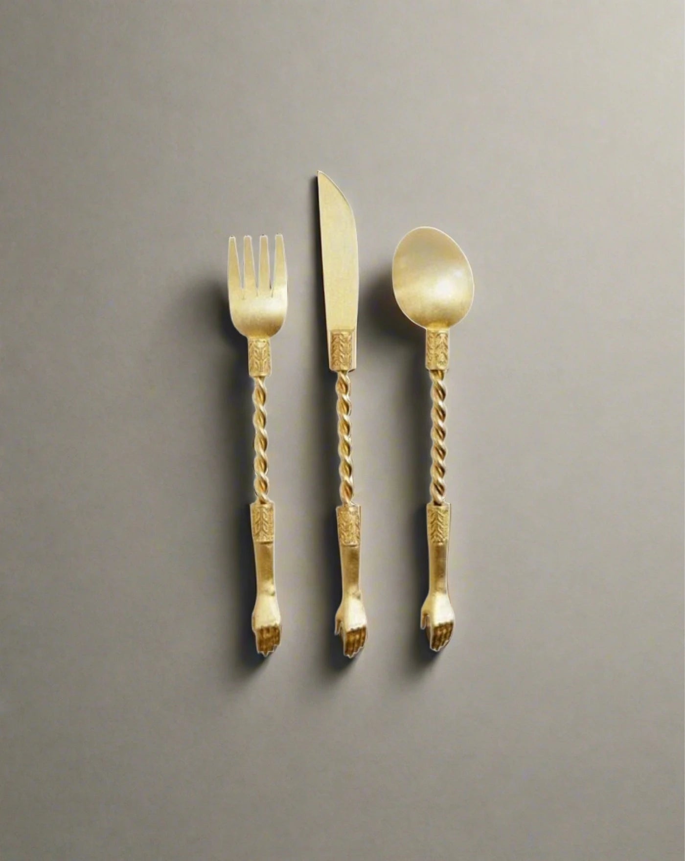 The Hand Set by Natalia Criado includes a gold-colored fork, knife, and spoon with elegantly twisted handles. This handcrafted collection enhances any dining experience with luxurious sophistication, set against a plain grey background.