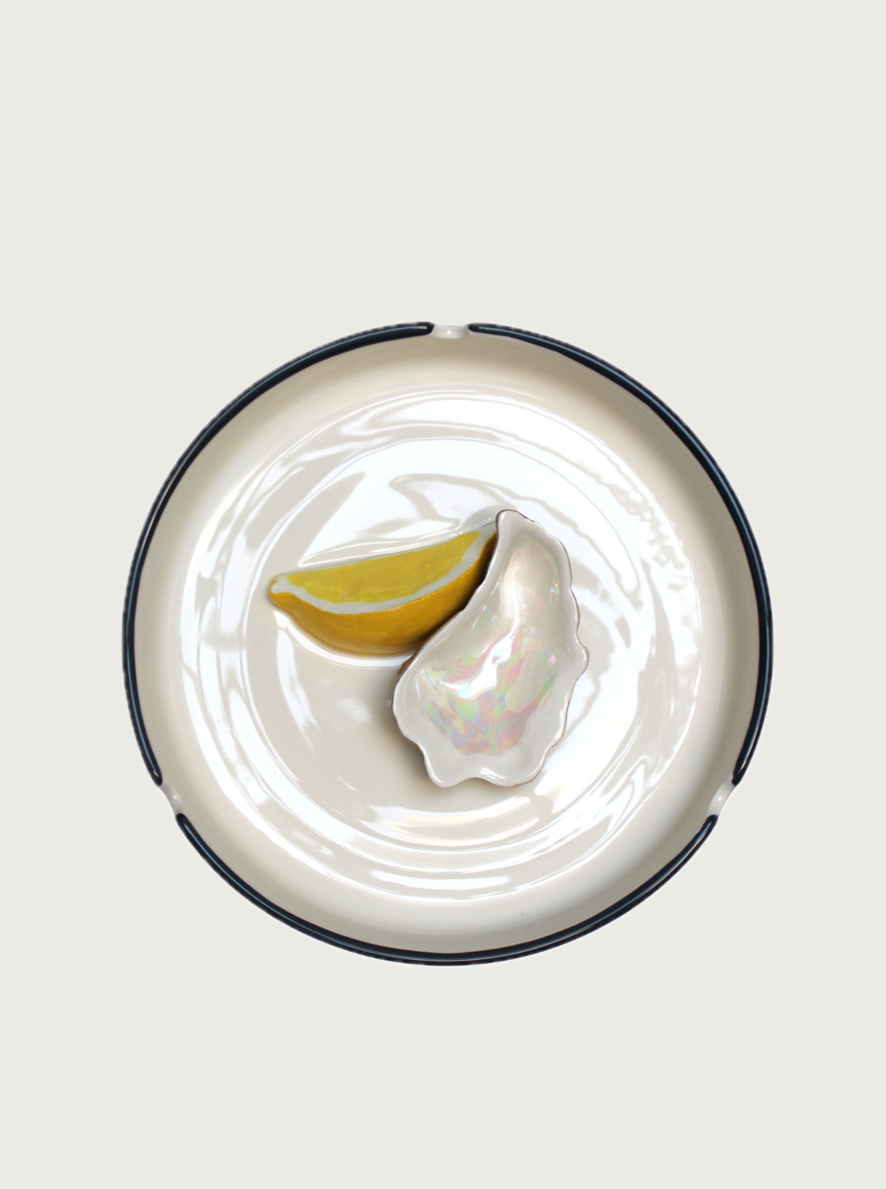 A pristine white plate with a black rim holds a shiny, iridescent Plaisir Solitaire Ashtray by Villa Arev and a slice of lemon, both centered in the middle. The scene evokes Mediterranean vibes reminiscent of Villa Arev, set against a solid white background.