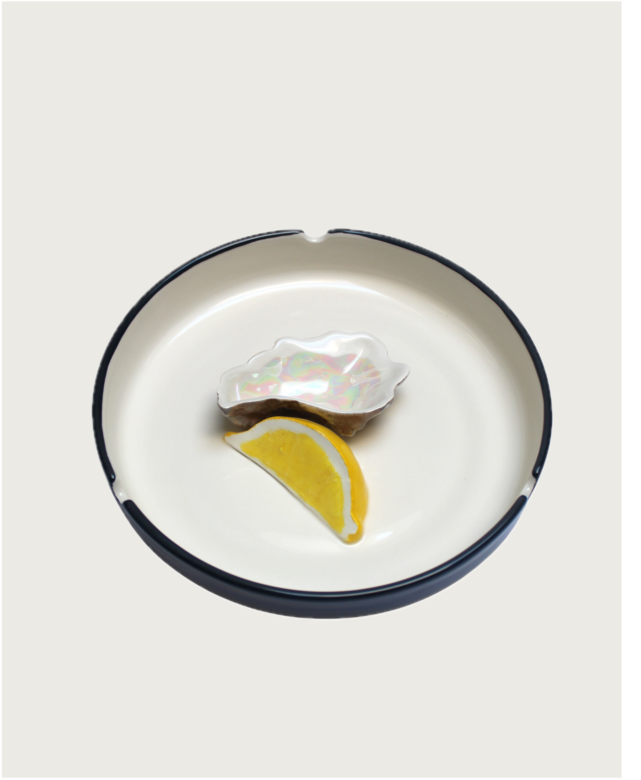 A Plaisir Solitaire Ashtray from Villa Arev with a decorative oyster shell and a lemon wedge made of ceramic, exudes summer vibes. The light-colored interior juxtaposed with a dark rim enhances its charm. This piece radiates warmth, reminiscent of sunlit days by the sea.