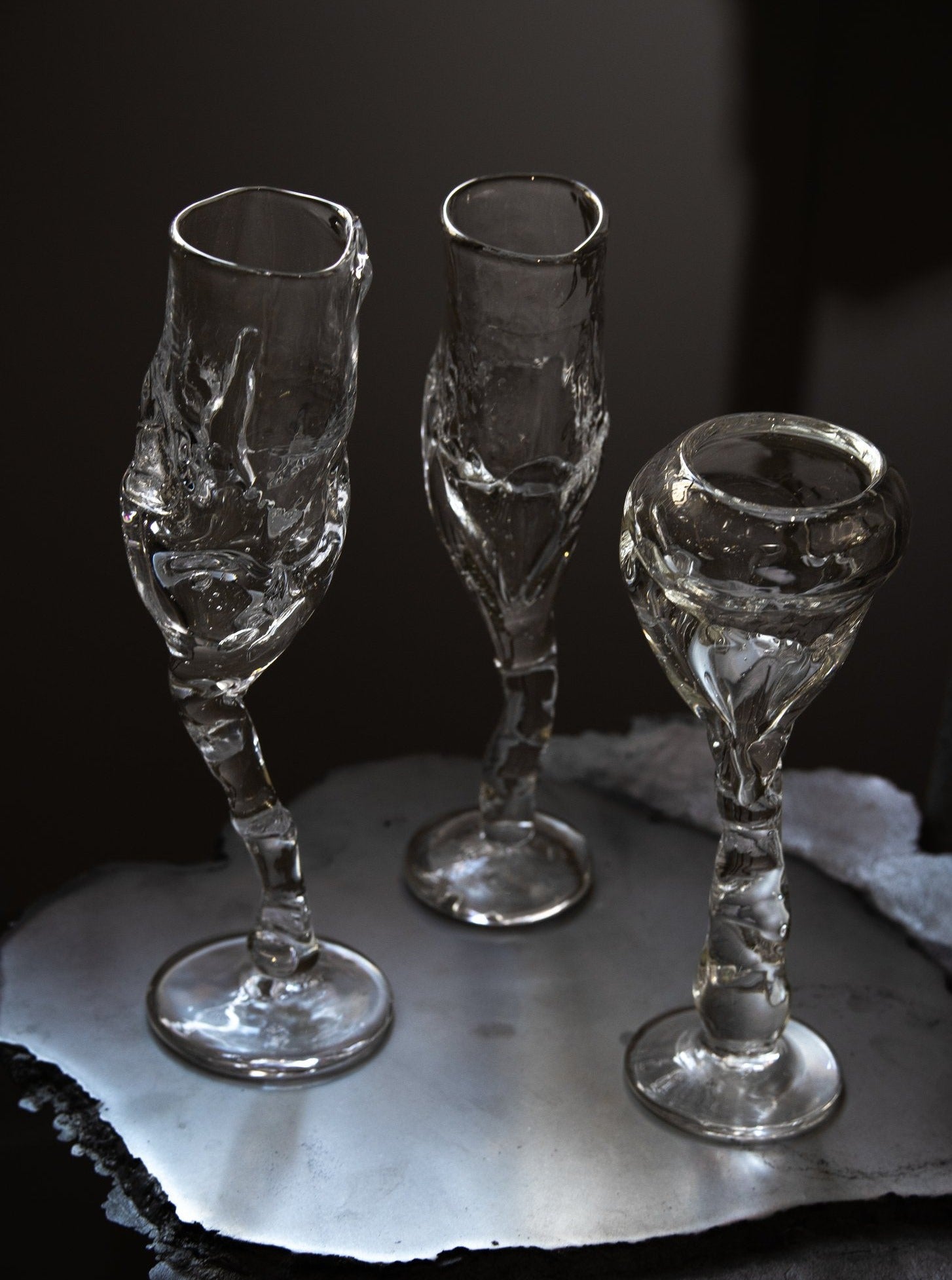 Three Flute - Transparent champagne glasses, hand-crafted by master artisans at Szkło Studio with twisted stems and irregular rims, are placed on a reflective, uneven surface in dim lighting. The glass has a jagged, almost organic appearance, emphasizing the unique and creative design.
