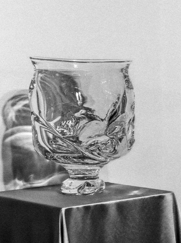 Black and white photo of an intricately designed Szkło Studio blown glass vase with foot, featuring floral patterns, standing on a draped pedestal.