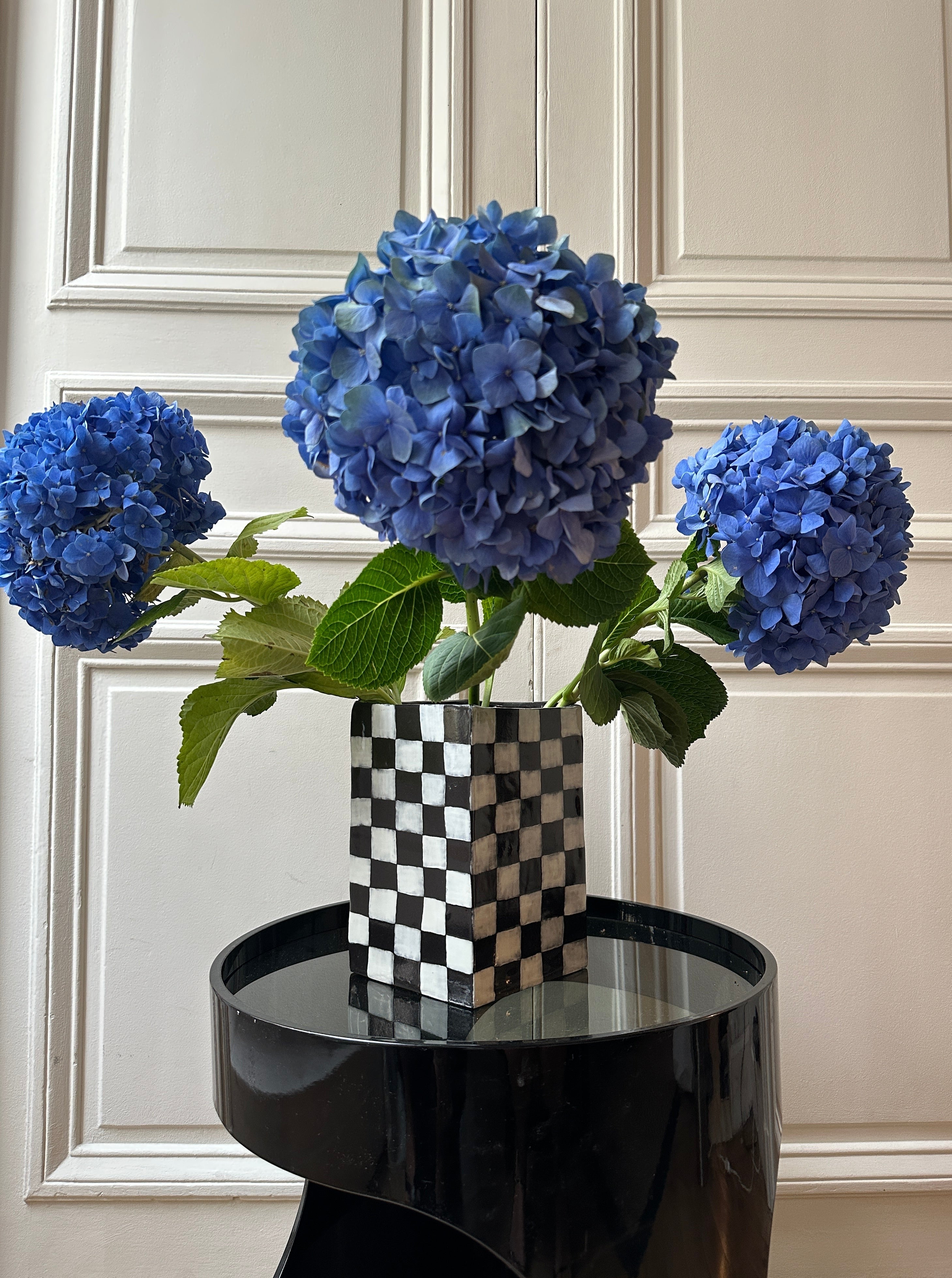 Three vibrant blue hydrangea blooms in a burrata studio Checkerboard Vase, placed on a reflective black table against a white paneled wall.