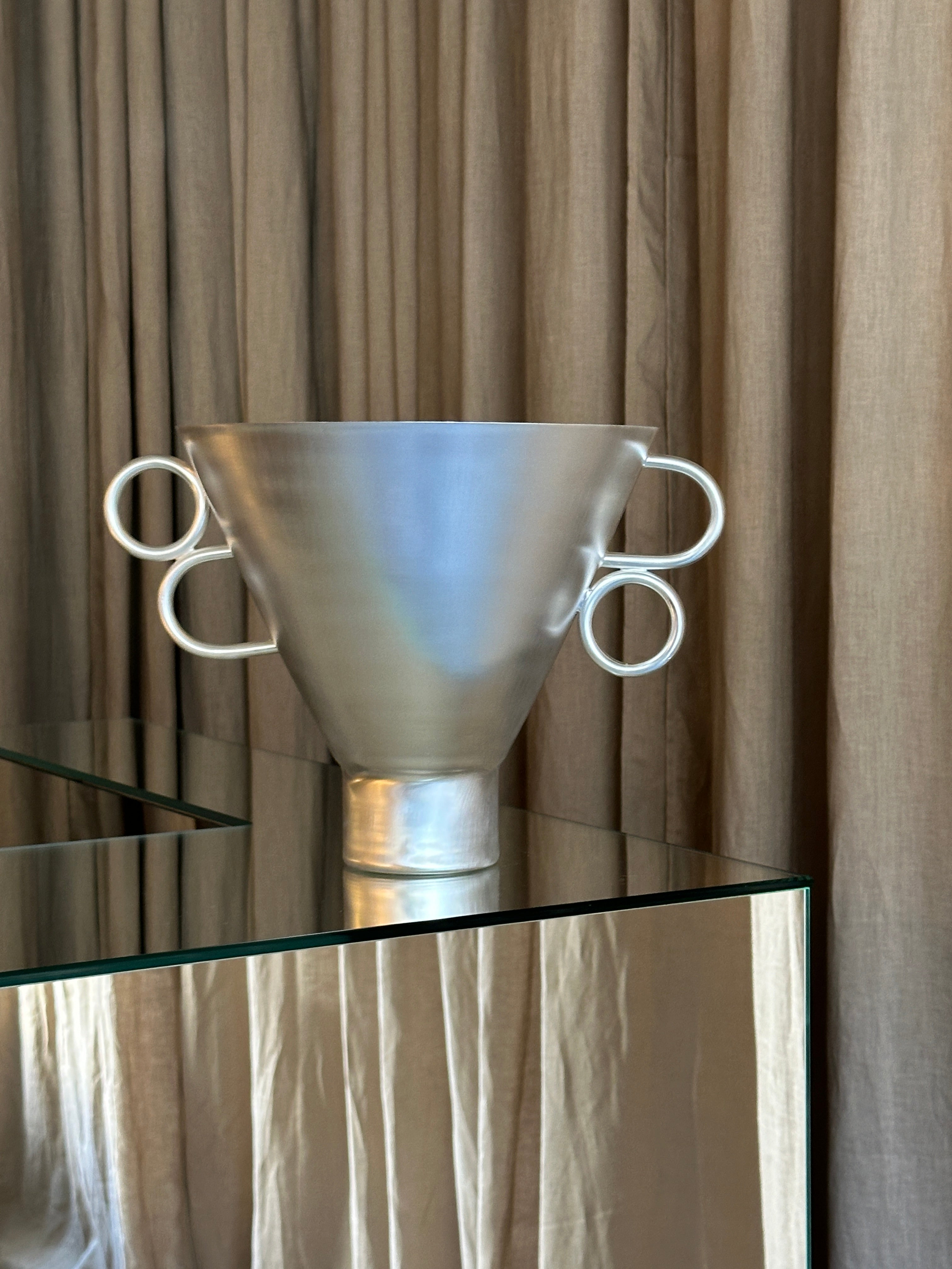 The Vaso Triangulare by Natalia Criado, a modern geometric vase with four circular side handles, sits on a glass table before beige curtains, adding a striking, sculptural touch to contemporary home decor.