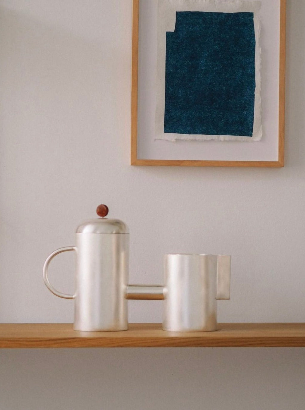 A minimalist Natalia Criado Oho silver teapot and mug featuring geometric handles sit on a wooden shelf, embodying contemporary design. A framed abstract artwork with a textured dark blue rectangle hangs on the white wall behind them, enhancing the minimalist aesthetic.