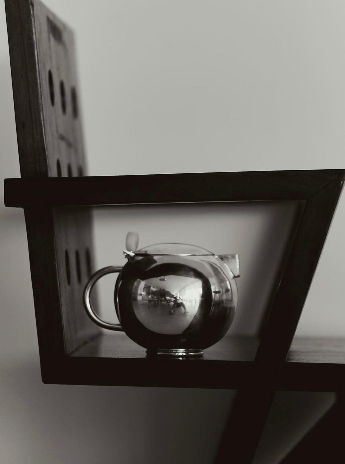 A monochrome image showing a clear, round Natalia Criado Teiera on a wooden shelf. The teapot reflects an inverted image of its surroundings during teatime gatherings.