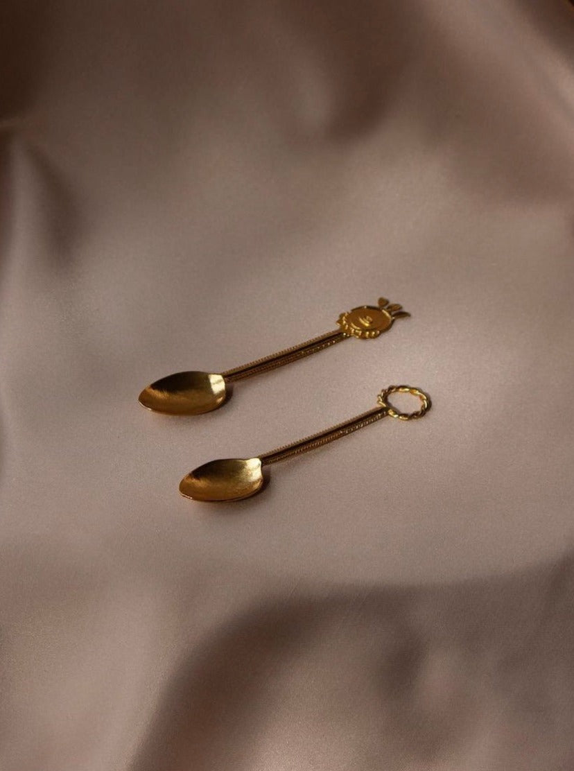 Two ornate, gold-colored Natalia Criado Wreaths Tea Spoons rest on silky taupe fabric. One has a floral design, the other an open circular handle. These handcrafted spoons beautifully reflect light, showcasing their metallic finish and minimalist aesthetic.