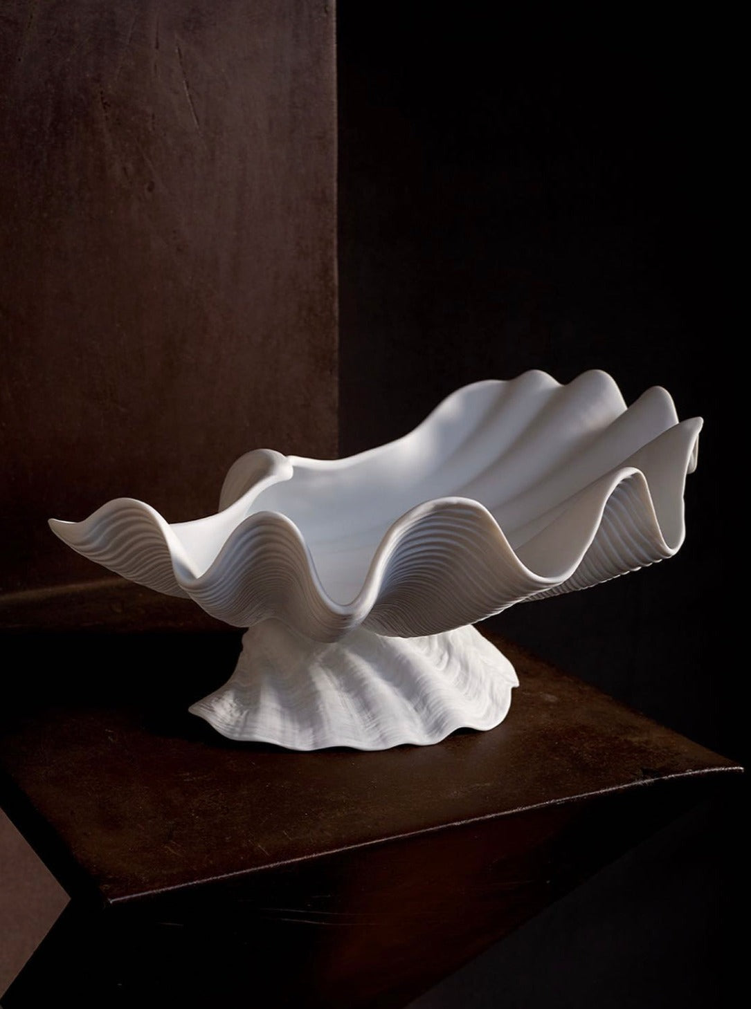 A sculptural white Neptune Bowl X-Large from the LOBJET Atelier Collection with undulating edges is displayed on a dark, rustic wooden table, against a dark backdrop, emphasizing its unique, Wabi-Sabi-inspired design.