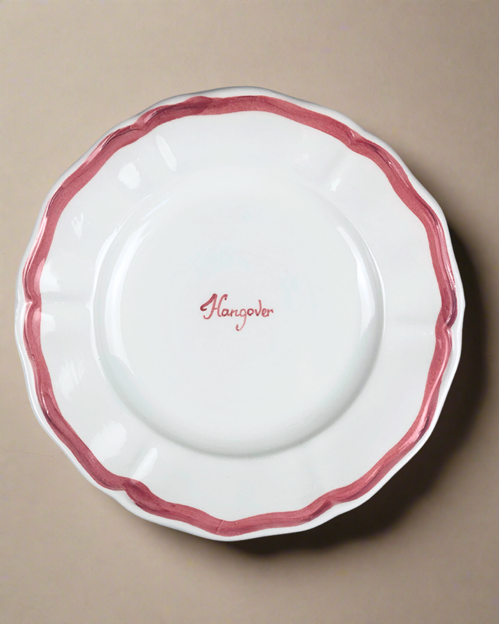 The "Hangover" Fil Rouge Plate by Musae Studio is a hand-painted white ceramic dish from Cilento, Italy. It features a wavy red rim and the word "Hangover" elegantly scripted in the center against a simple beige background, enhancing its artistic appeal.