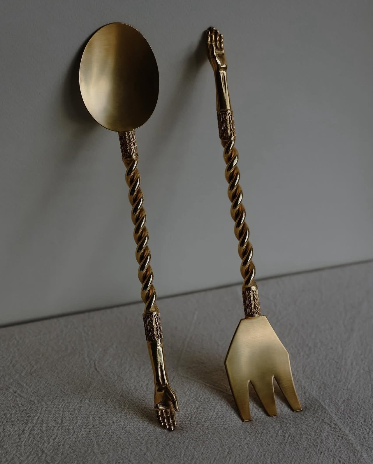 The Natalia Criado Hand Server Set features a gold-colored brass spoon and fork with twisted handles ending in sculpted hands, displayed on a textured gray surface—an ideal unique housewarming gift.