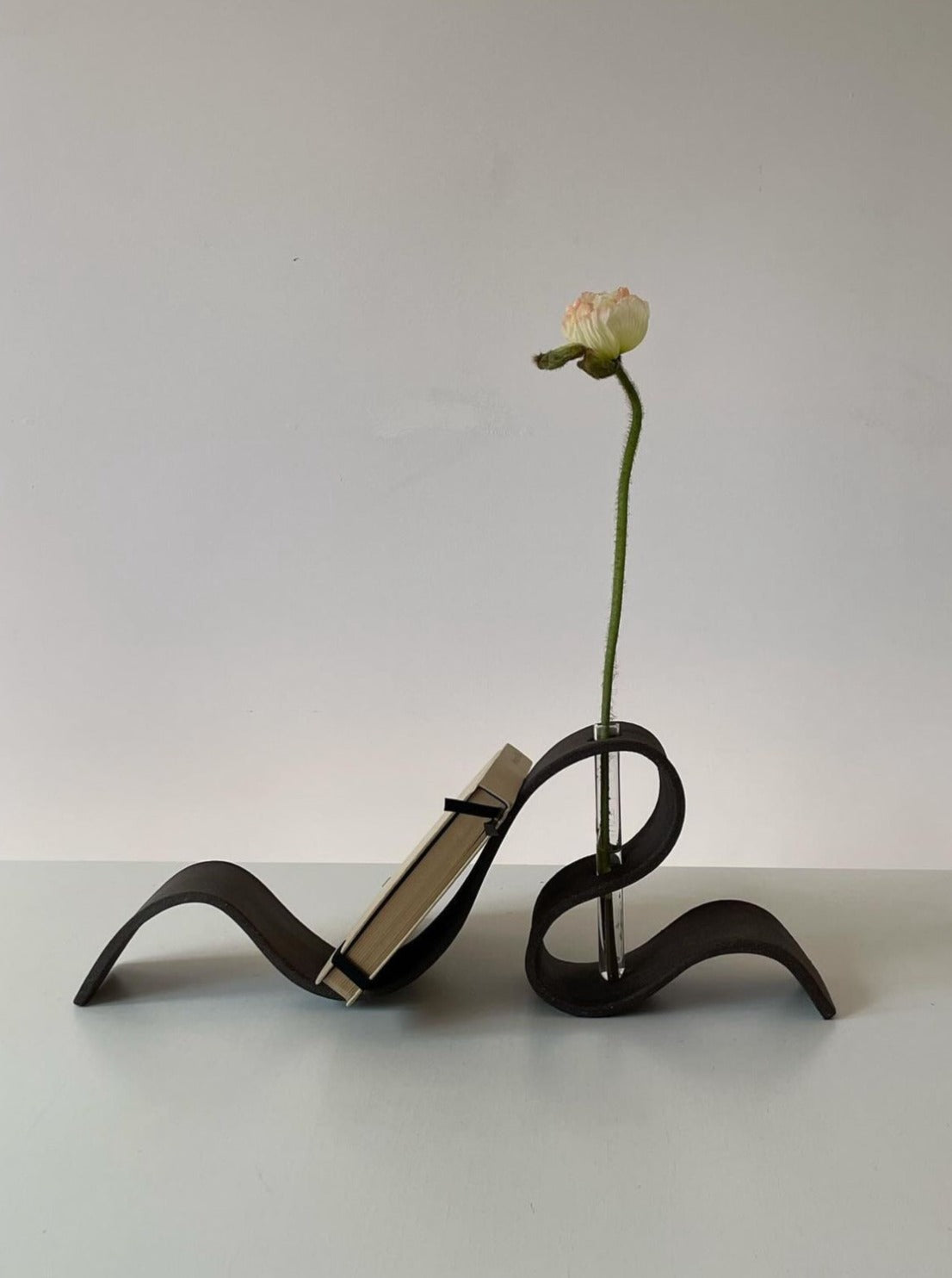 A minimalist sculpture features fluid, undulating forms holding an open book holder on the left and a single white flower with a thin, green stem in LESS's Ribbon Vase in Black on the right. The sculpture sits on a plain surface against a light grey background.