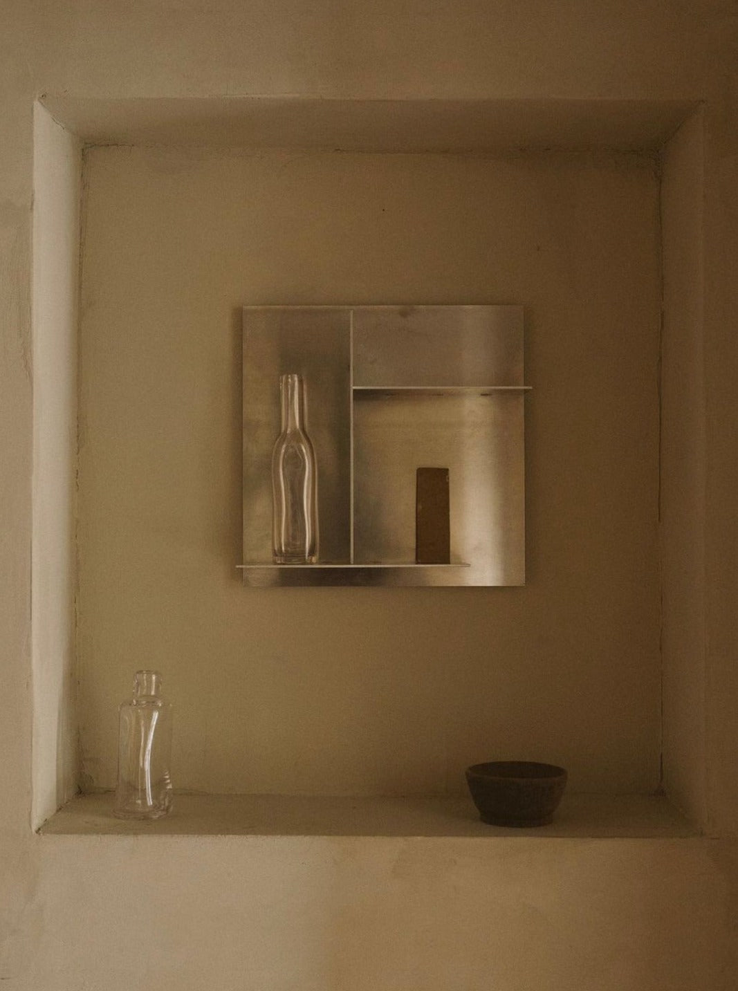 A minimalist art display featuring three objects: a metal panel with cut-out shapes of a bottle and narrow rectangle, a transparent glass bottle, and a small ceramic bowl by FRAMA, all positioned.