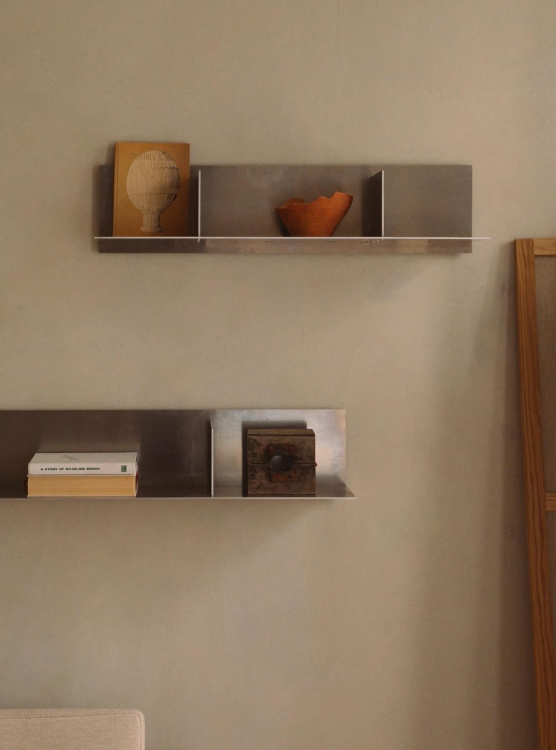 A minimalist room features floating FRAMA Rivet Shelf | Aluminum | Small on a neutral wall. The top shelf holds a small sculpture and a wicker vase, while the lower shelf displays a few books and a small framed artwork. A touch of raw aluminum decor accents the space, with the corner of a beige couch visible at the bottom left.