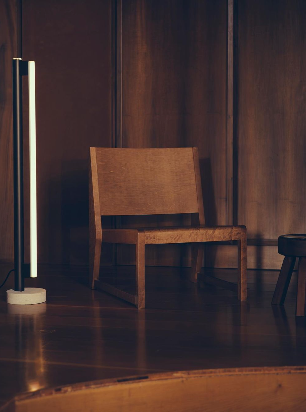 A modern minimalist room with a wooden Easy Chair 01 | Warm Brown Birch by FRAMA featuring a brown oiled birch finish and a tall, slim floor lamp. The design aesthetic is simple, with dark wood paneling on the wall. A small black stool or side table is partially visible in the background.