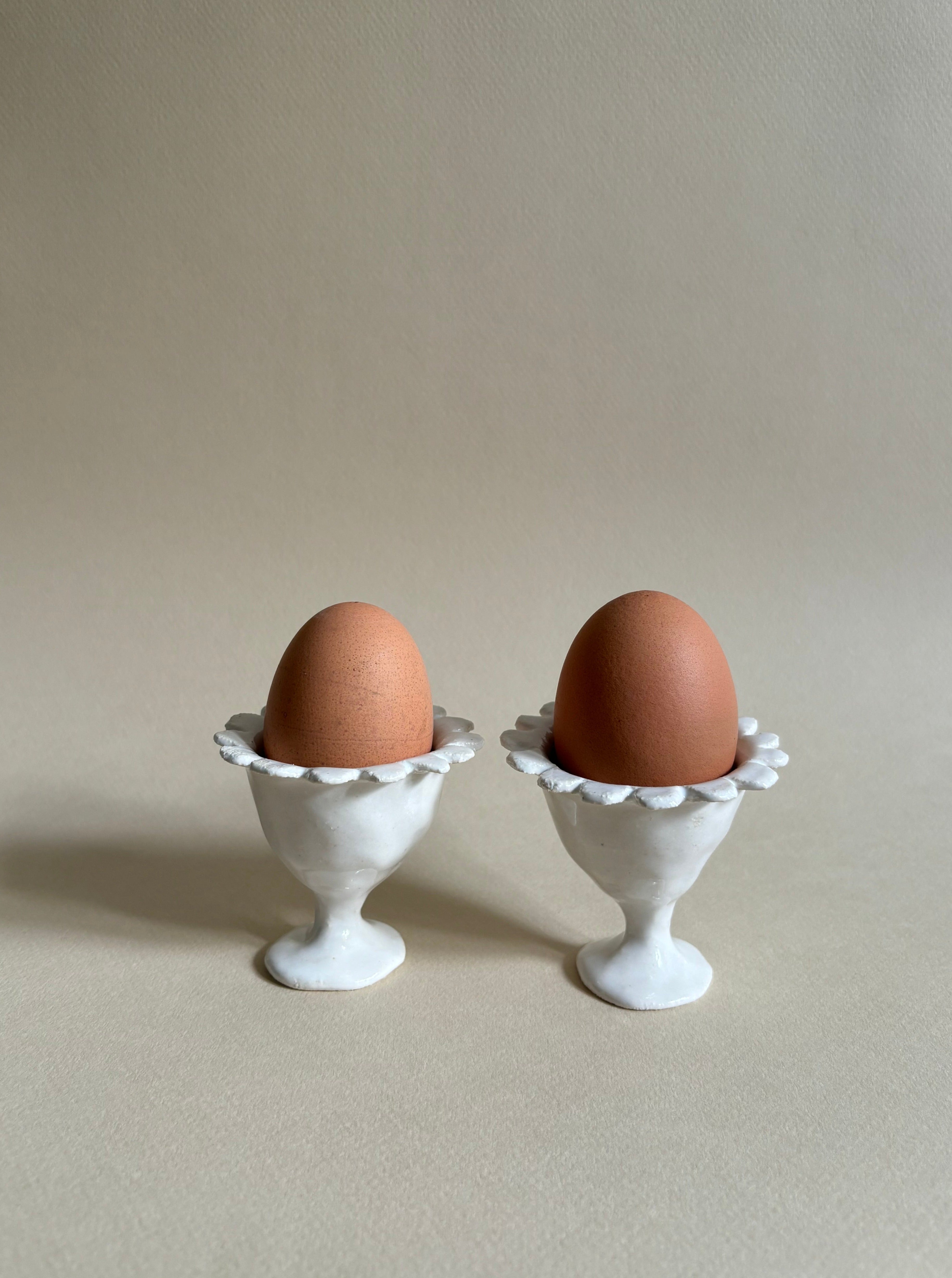 Two brown eggs are placed in white, ornate egg cups. The "Rosetta" Egg Cups, crafted by Studio Beurre from white clay with a shiny white glaze, add an artisanal touch. The background is a plain light beige, providing a simple and neutral setting that emphasizes the eggs and their holders. Soft lighting creates gentle shadows.