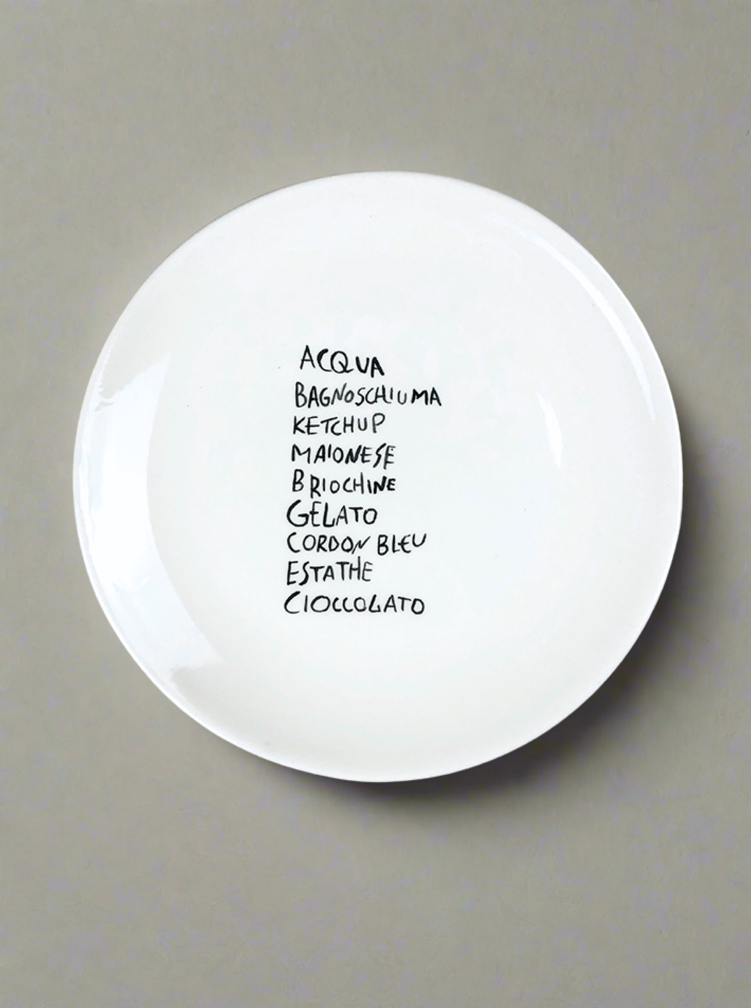 The "Tommy's Essentials" Plate by Musae Studio features a handwritten list of Italian words and sits on a gray background, adding a playful touch to elegant dining and making for unique tableware.