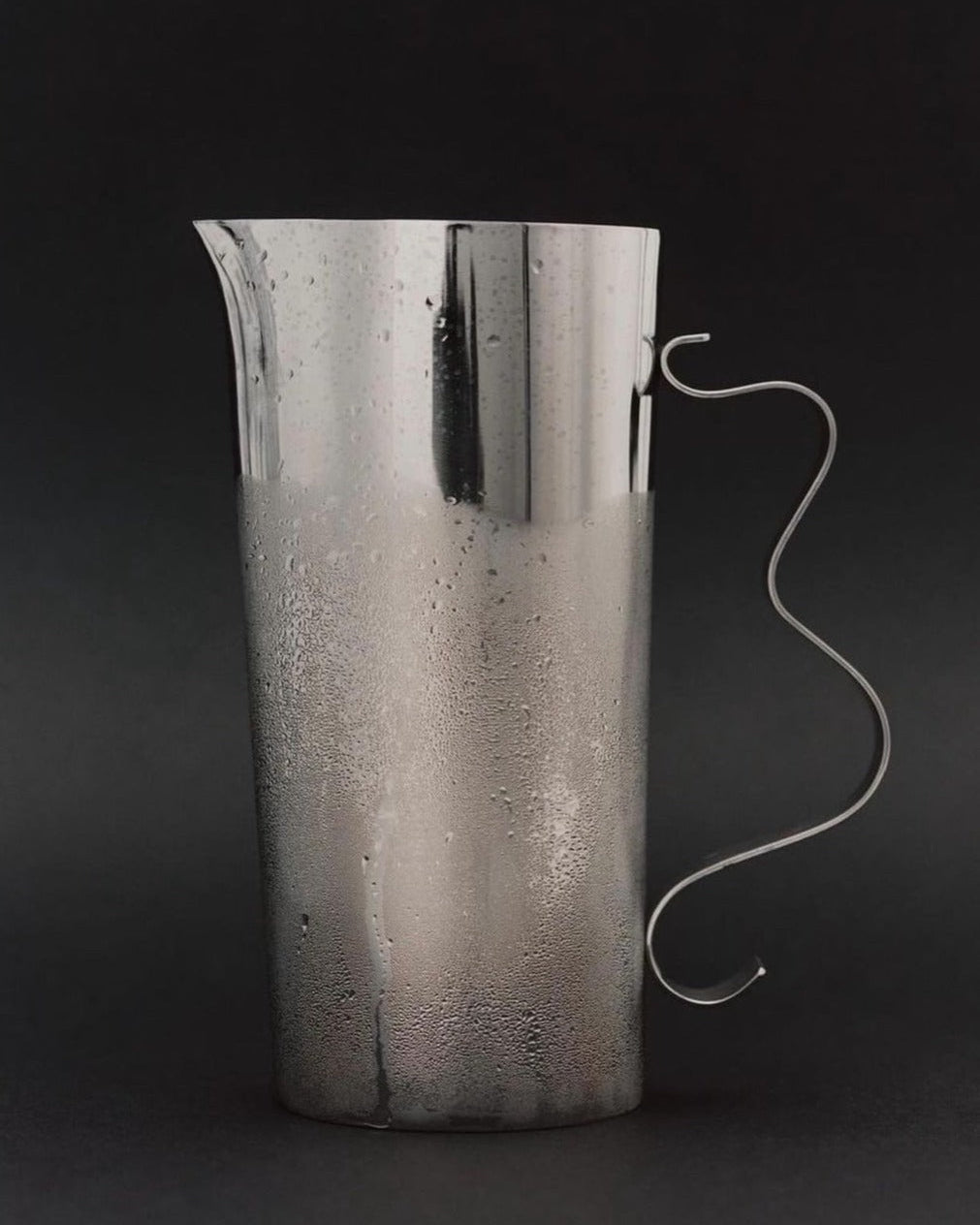 A tall, sleek stainless steel Squiggle Pitcher by Sophie Lou Jacobsen with a unique, curvy handle is shown against a dark background. Condensation droplets are visible on the pitcher’s surface, indicating it is cold. The design is simple and modern, making it an elegant counterpart to any Squiggle Tray or decorative vase.