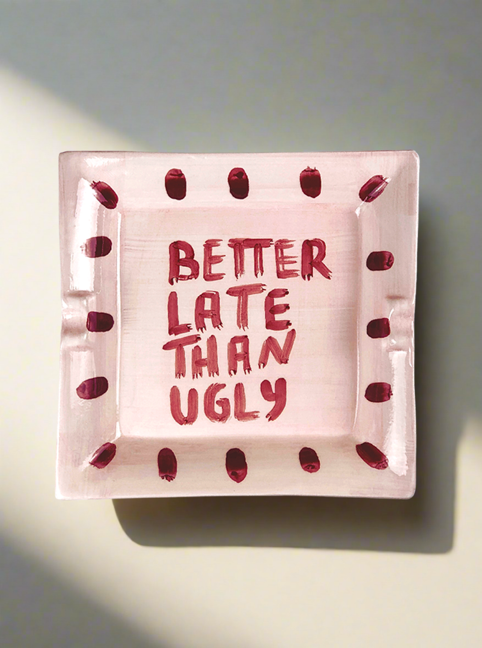 The "Better late than ugly" Ashtray by Musae Studio is a square pink ceramic piece with red dot edges, featuring the playful phrase "BETTER LATE THAN UGLY" in red at its center, making it a fun and functional catchall.
